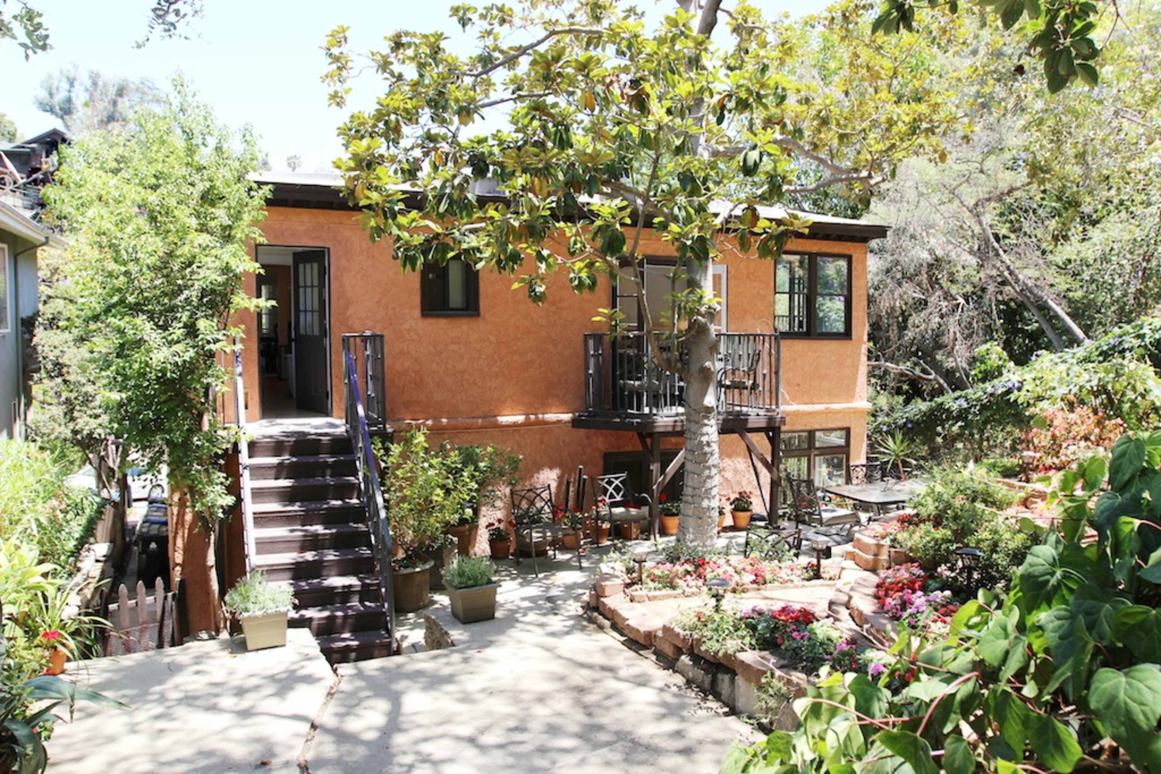 A two-story house with orange walls and black railings is surrounded by a garden featuring potted plants and flowers.