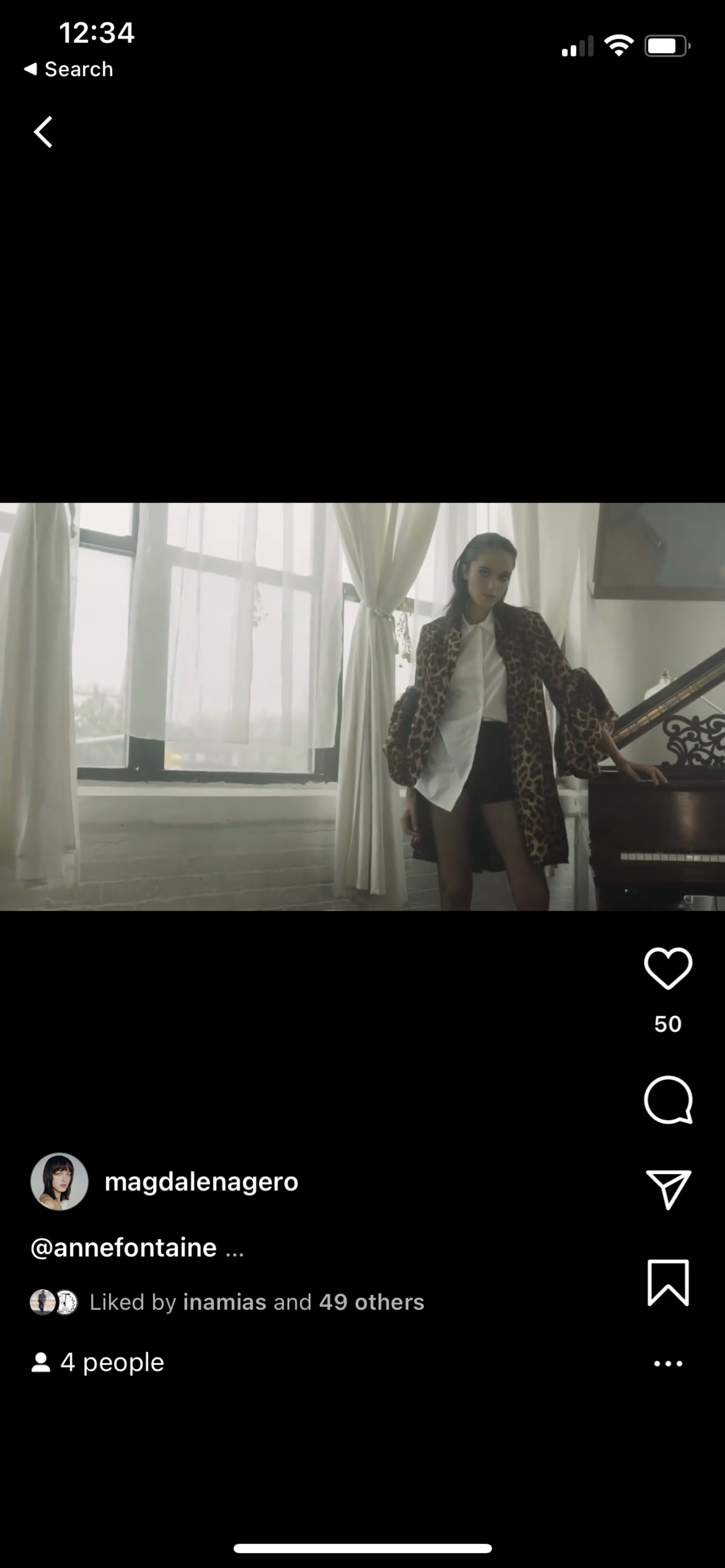 A person stands next to a grand piano in a room with large windows and light-colored drapes.