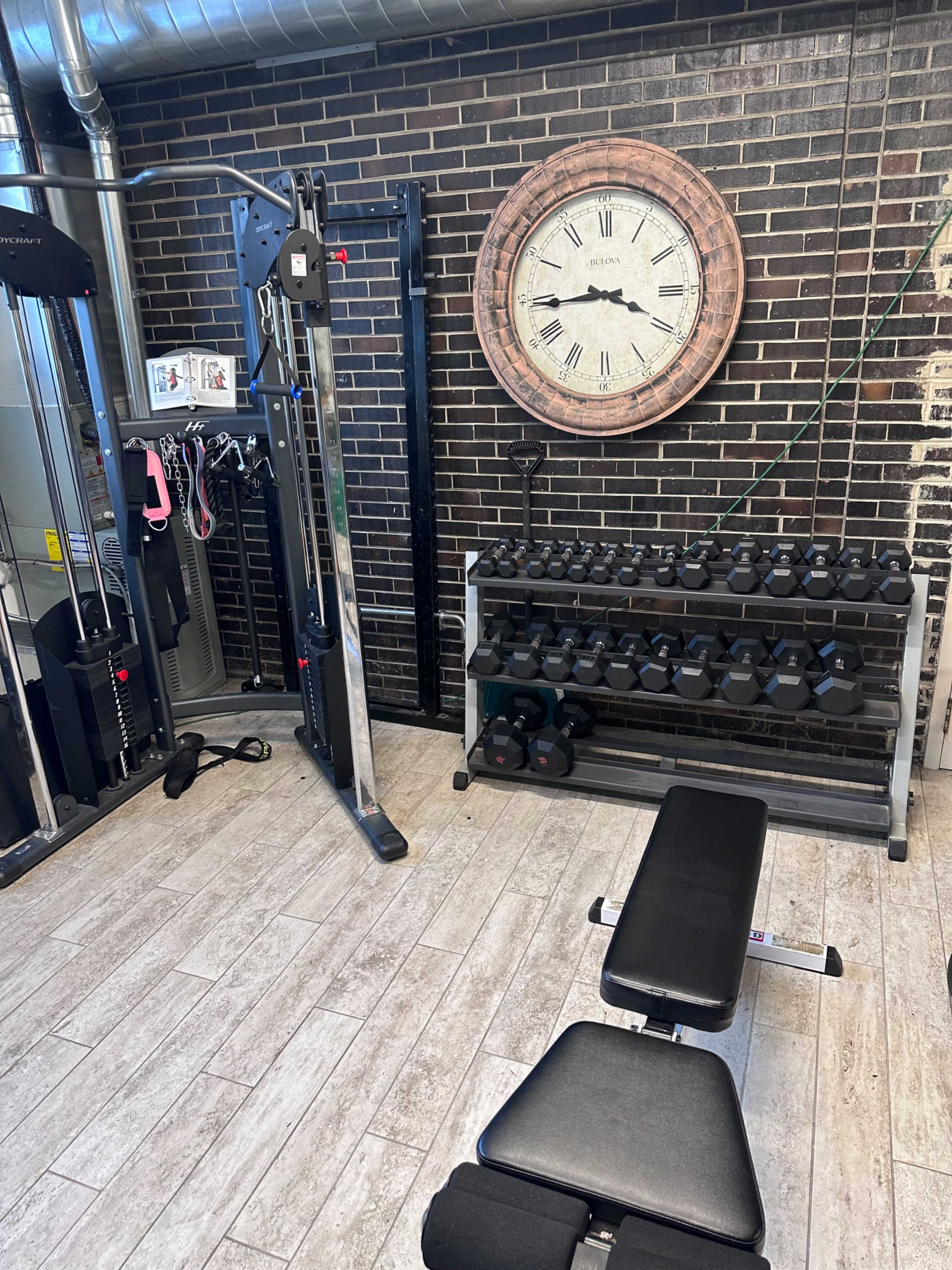 A home gym area with a weight bench, dumbbell rack, and a cable machine against a brick wall, complemented by a large wall clock.