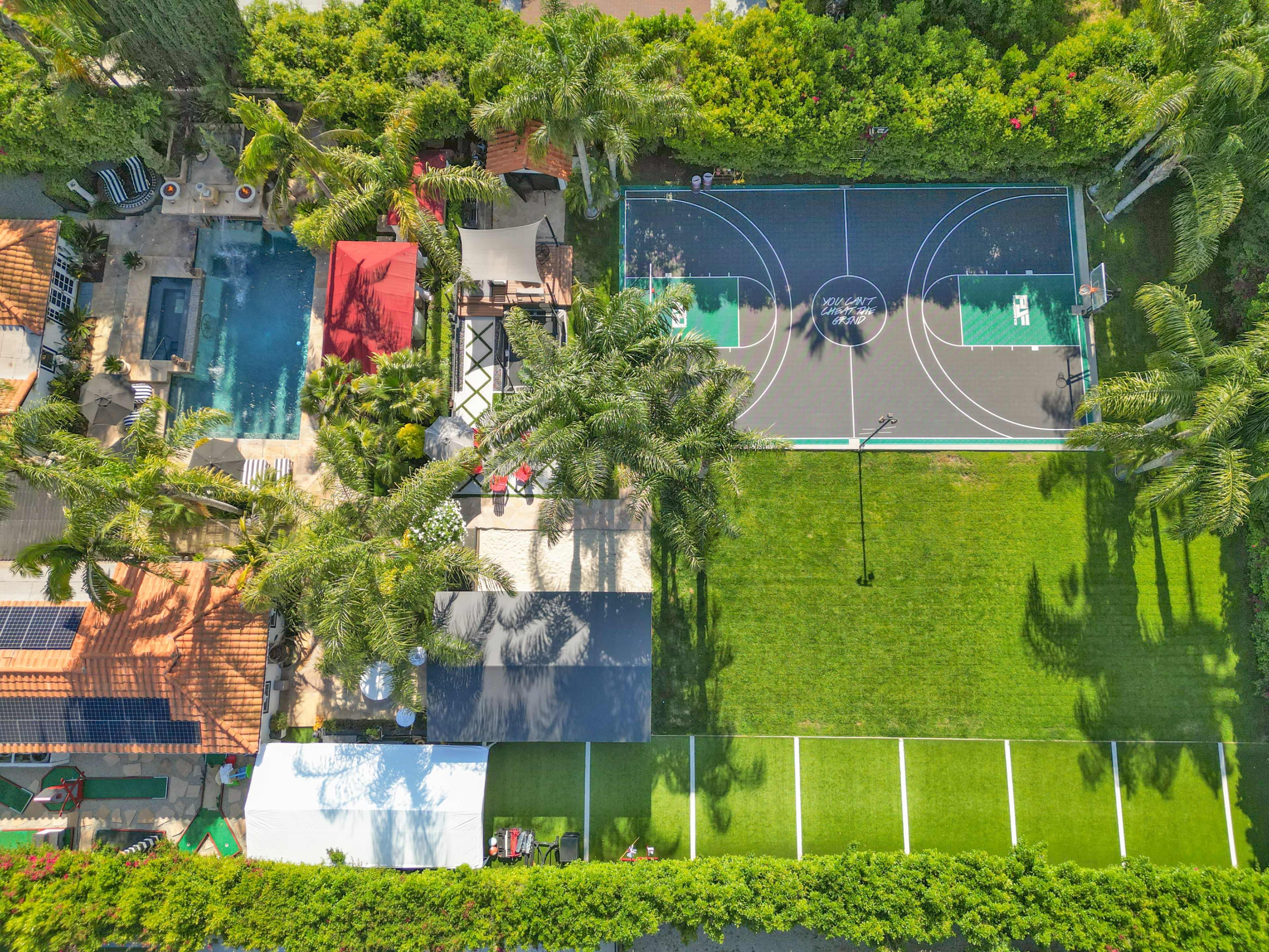 A spacious backyard featuring a basketball court, a swimming pool, and various outdoor seating areas surrounded by palm trees.