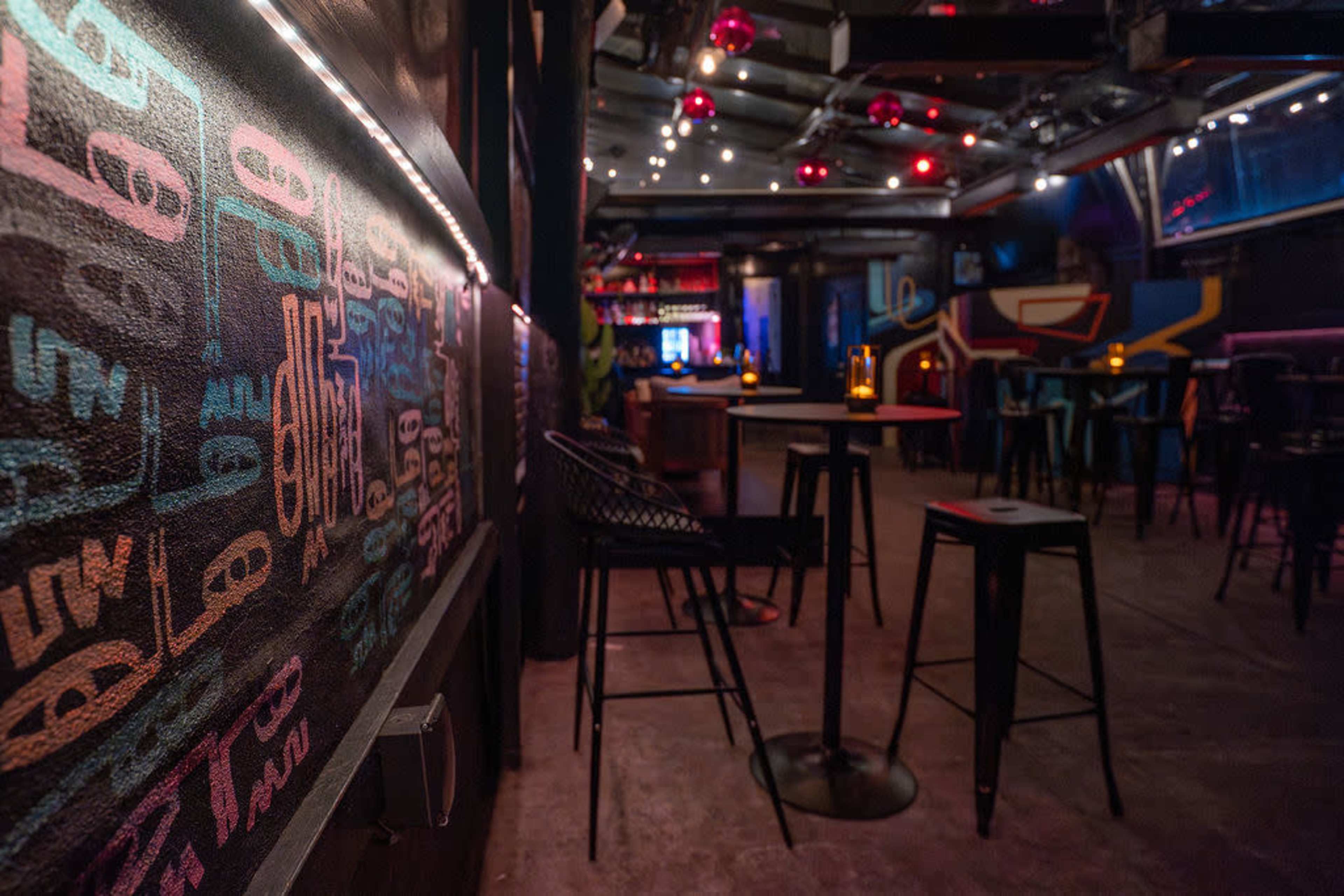 The image shows the interior of a bar with dim lighting, featuring graffiti on the walls, tall tables, and scattered chairs.