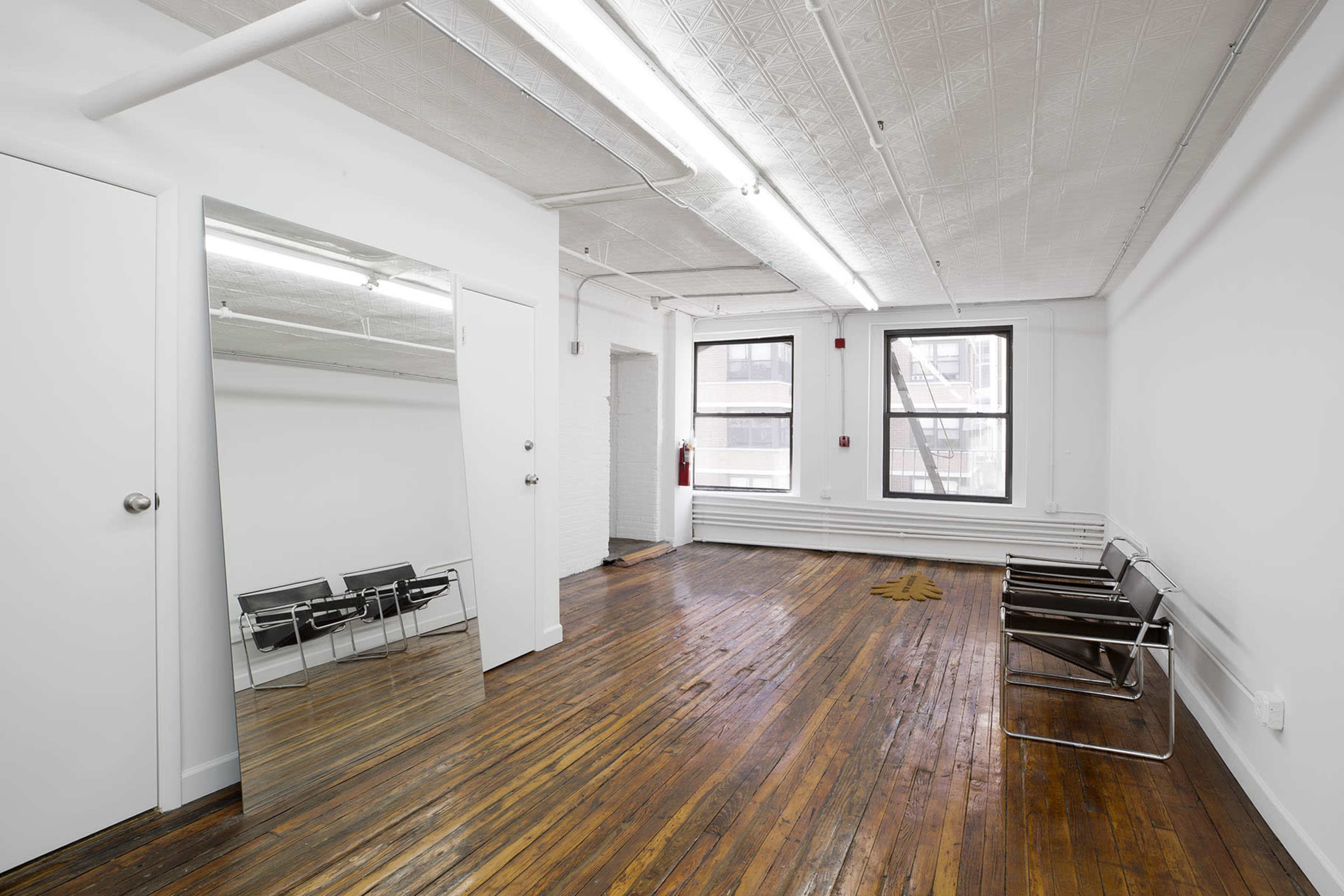 The image shows a spacious, empty room with hardwood floors, a large mirror on one wall, and two black chairs placed opposite a window.