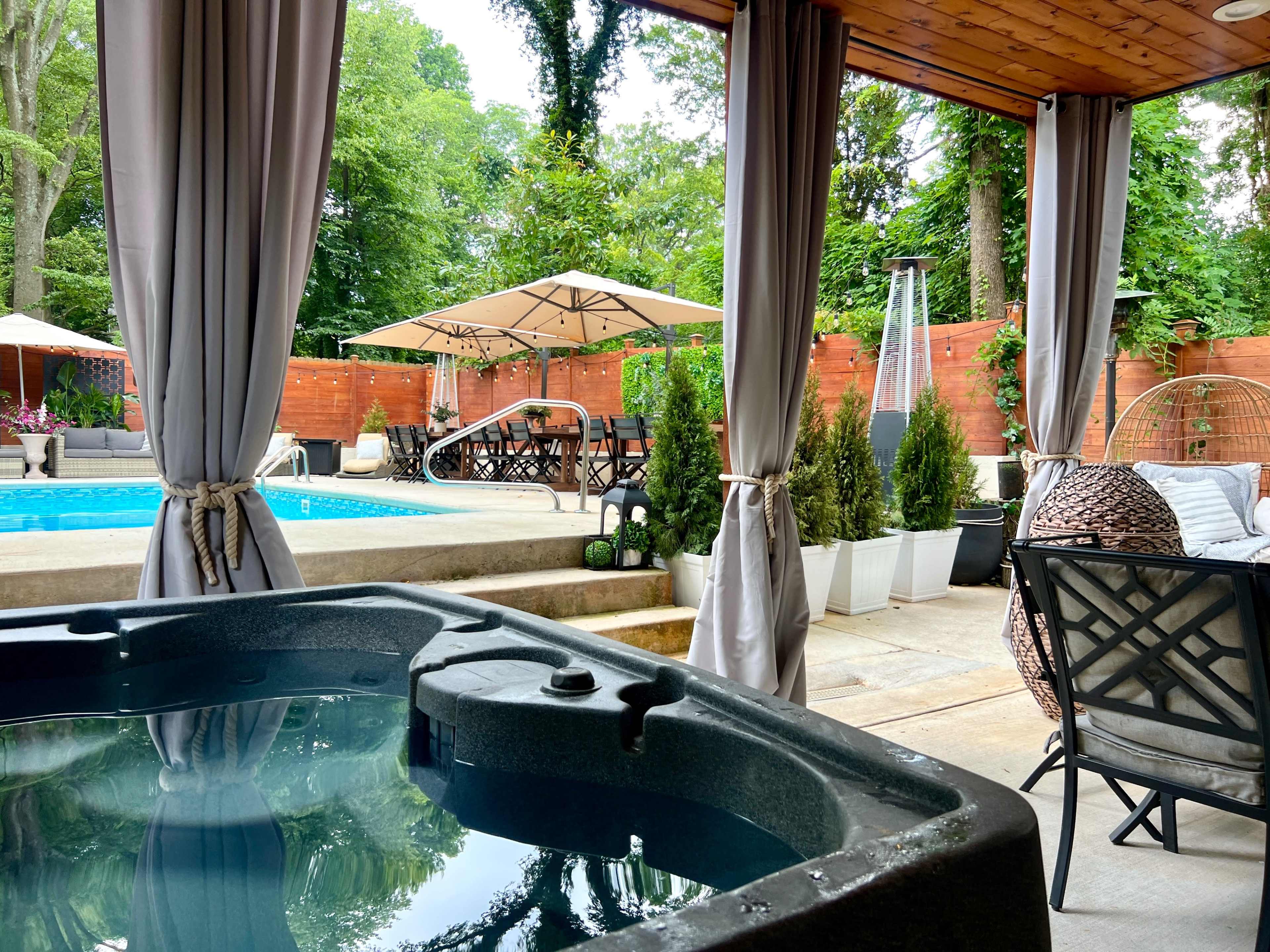 A backyard scene with a hot tub in the foreground, a swimming pool in the background, and various outdoor furniture and greenery.