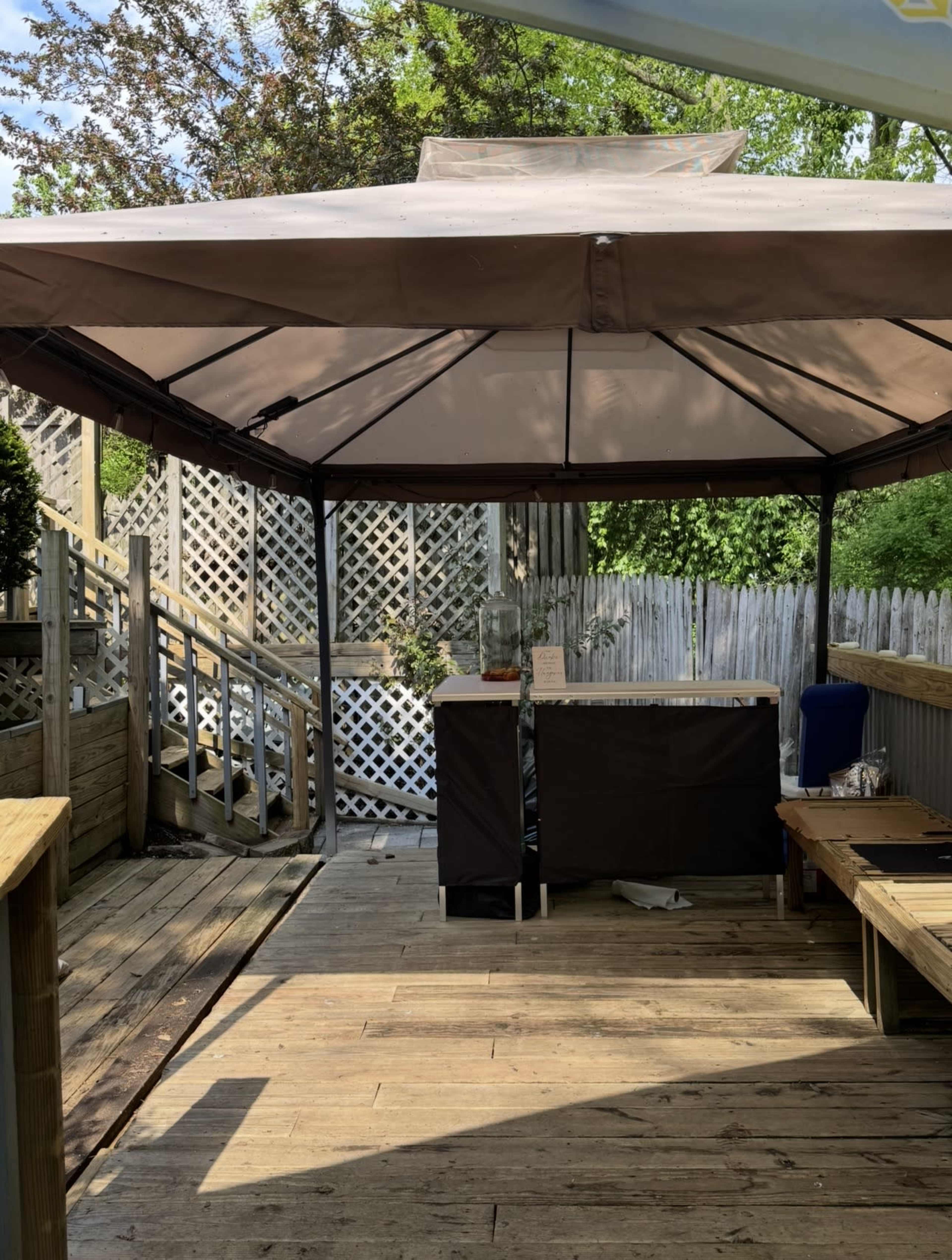 The image shows a wooden deck area with a gazebo, a bar setup, and a staircase leading to a lattice-fenced area with greenery in the background.