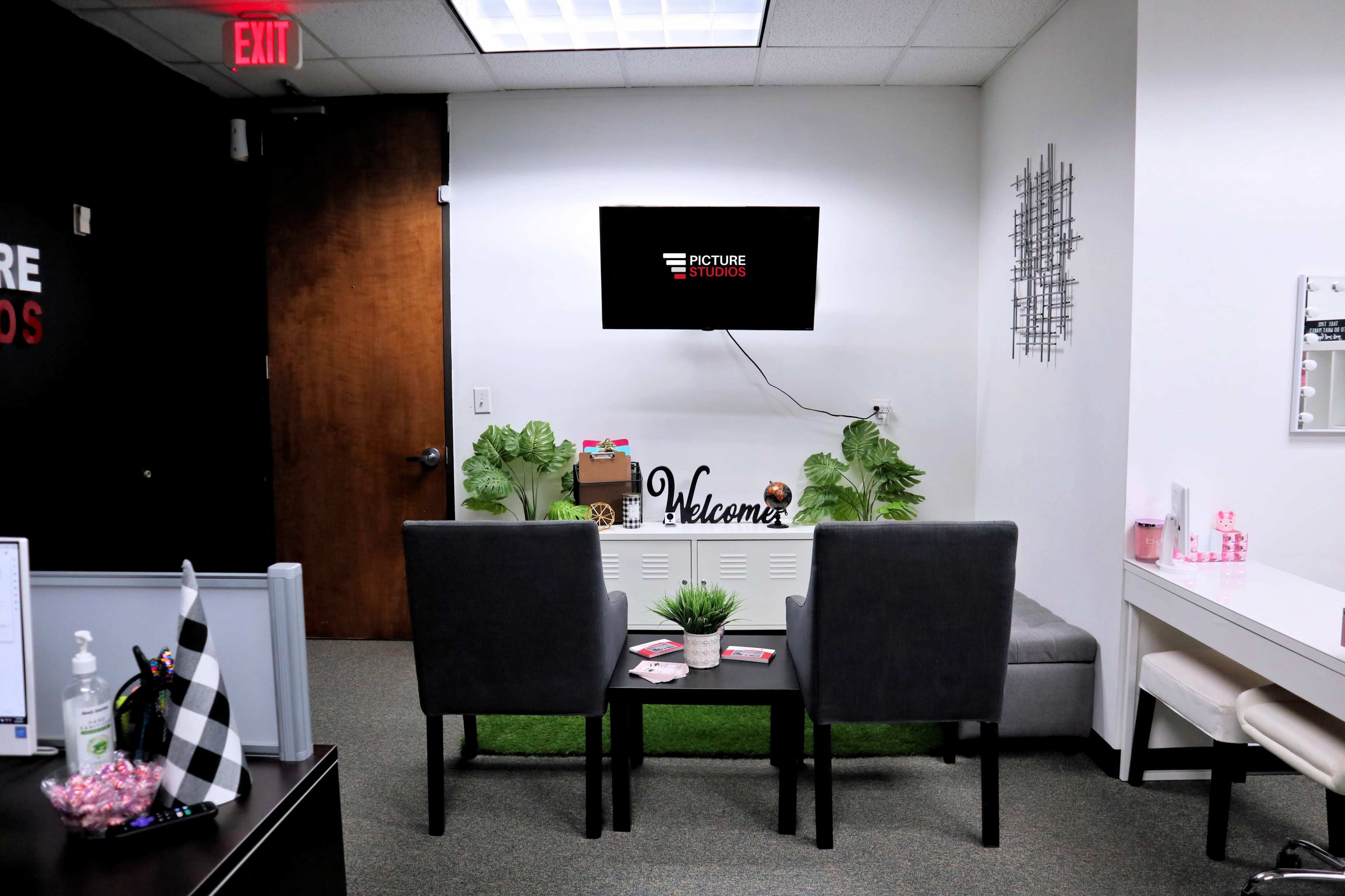 A small waiting area with two chairs, a table, and a television mounted on the wall, featuring decorative plants and a welcome sign.