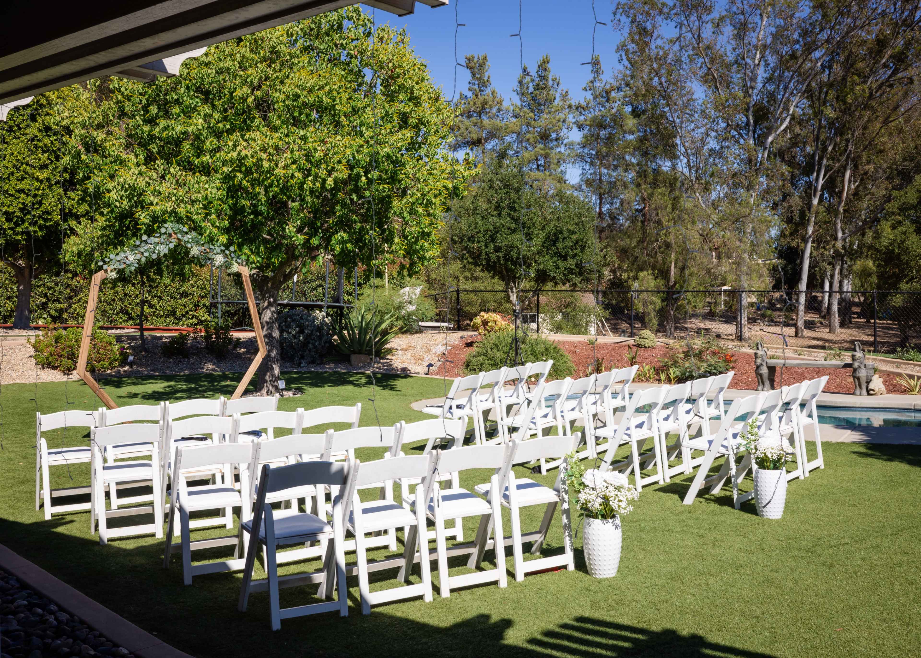 A set of white chairs is arranged in rows on a grassy area near a pool, with decorative flower pots positioned at the ends of the rows.