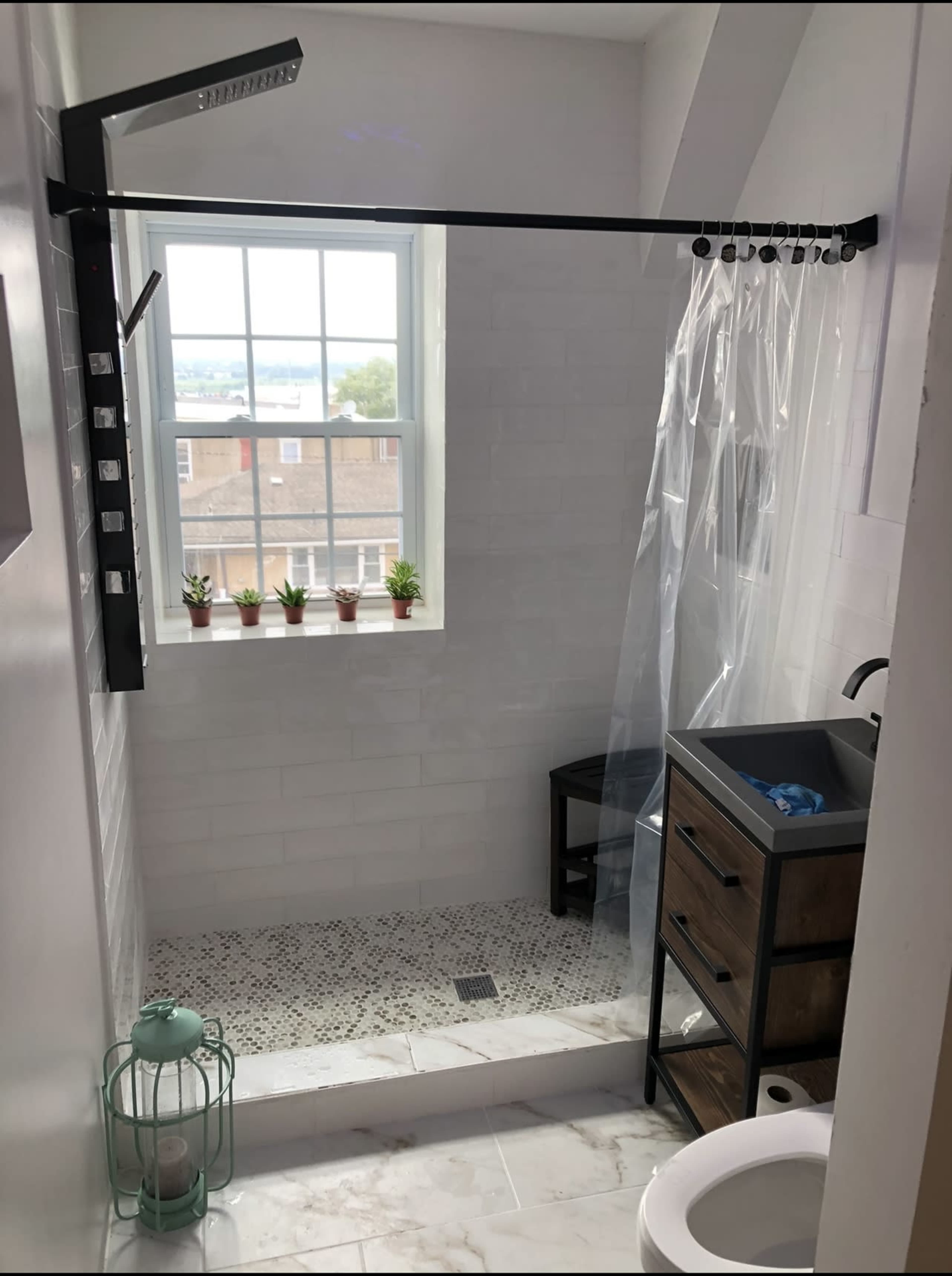 A modern bathroom with a walk-in shower featuring a clear curtain, a small sink cabinet, and a window with potted plants.