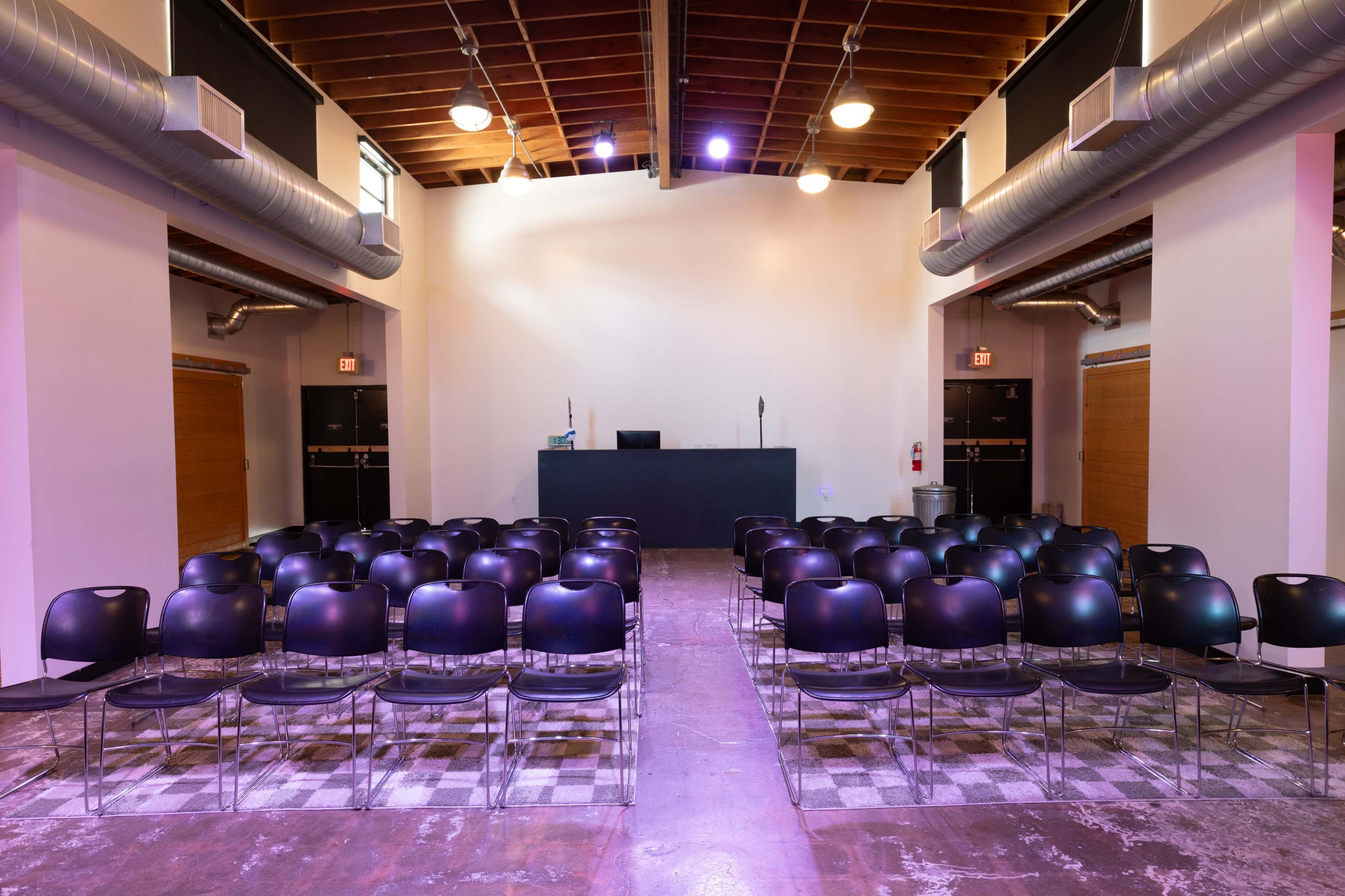The image shows a room configured with rows of black chairs facing a low stage or podium, with wooden doors on either side and ambient lighting.