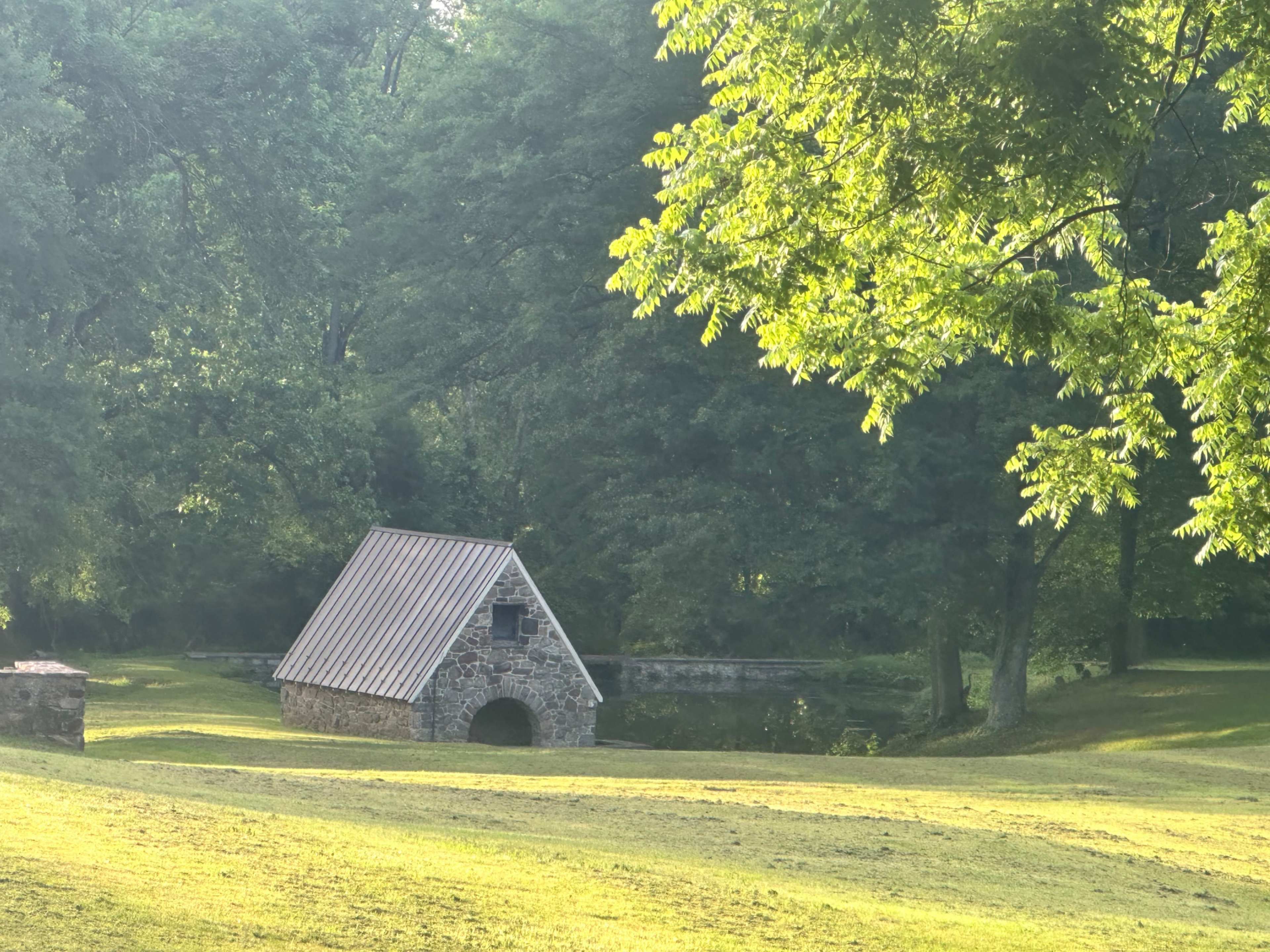 The image shows a small stone building with a sloped roof beside a pond, surrounded by lush greenery.