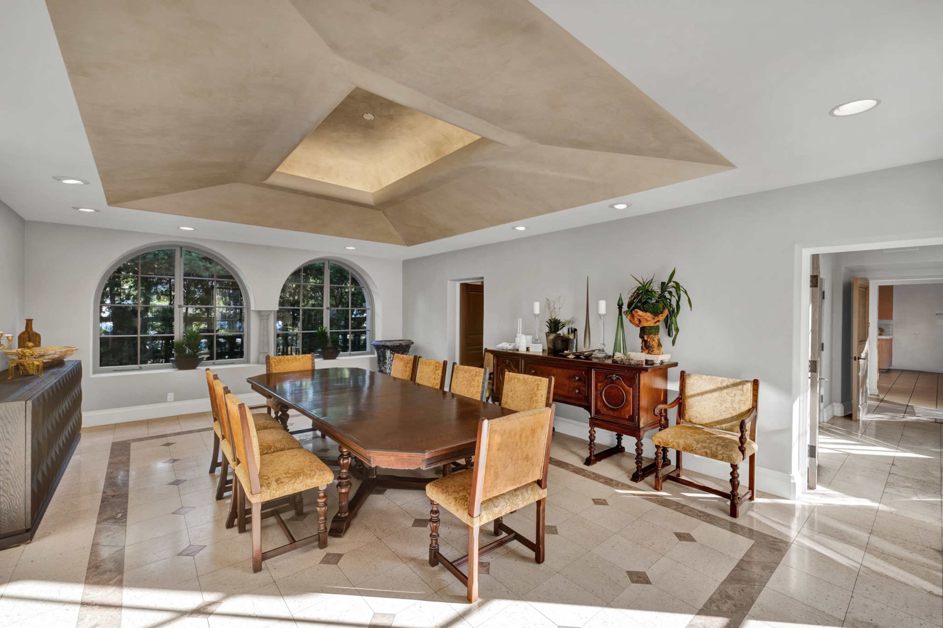 A spacious dining room features a large wooden table surrounded by upholstered chairs and windows that overlook a garden.
