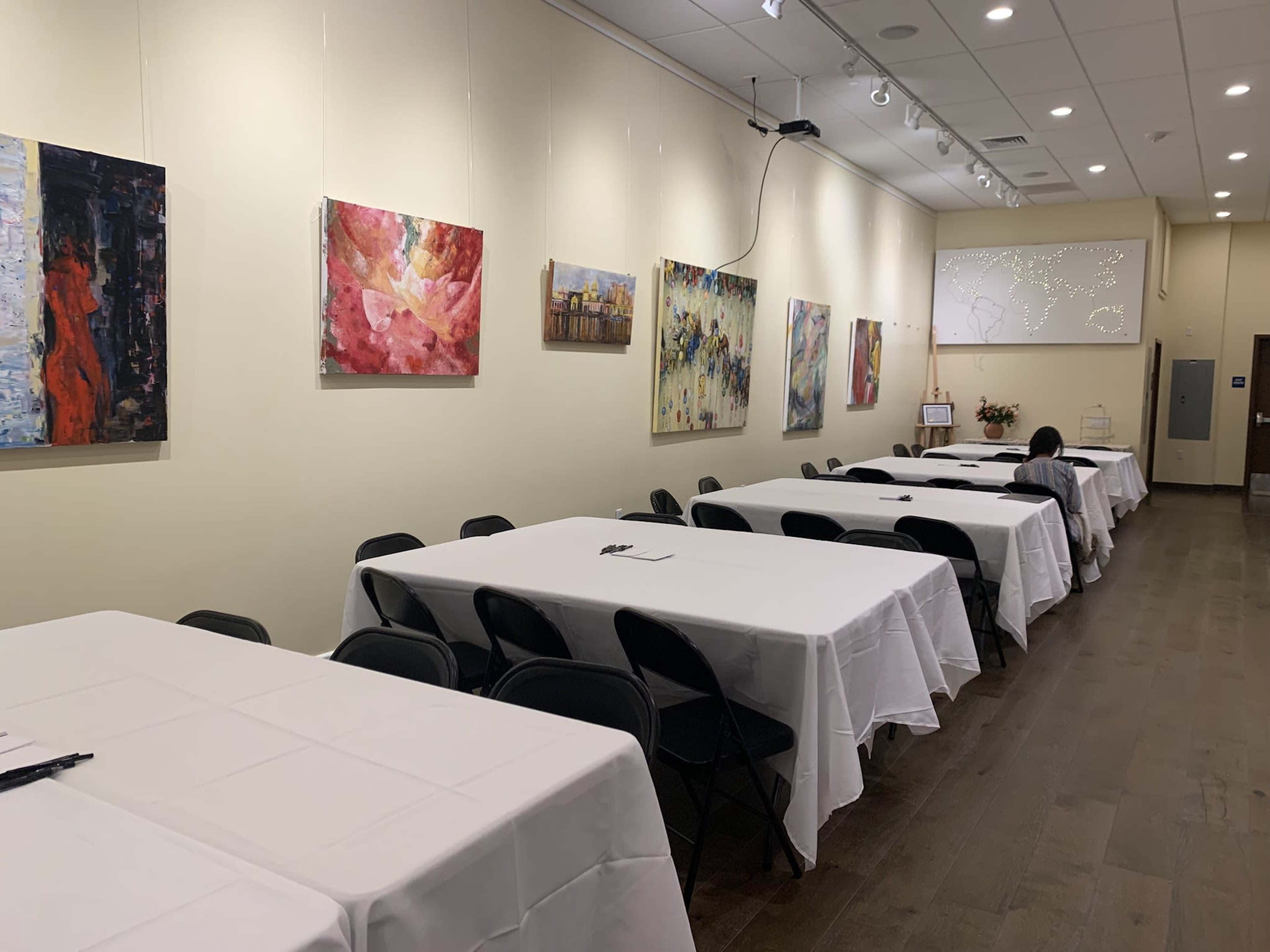 A room with several round tables covered in white tablecloths is lined with colorful abstract paintings on the walls.