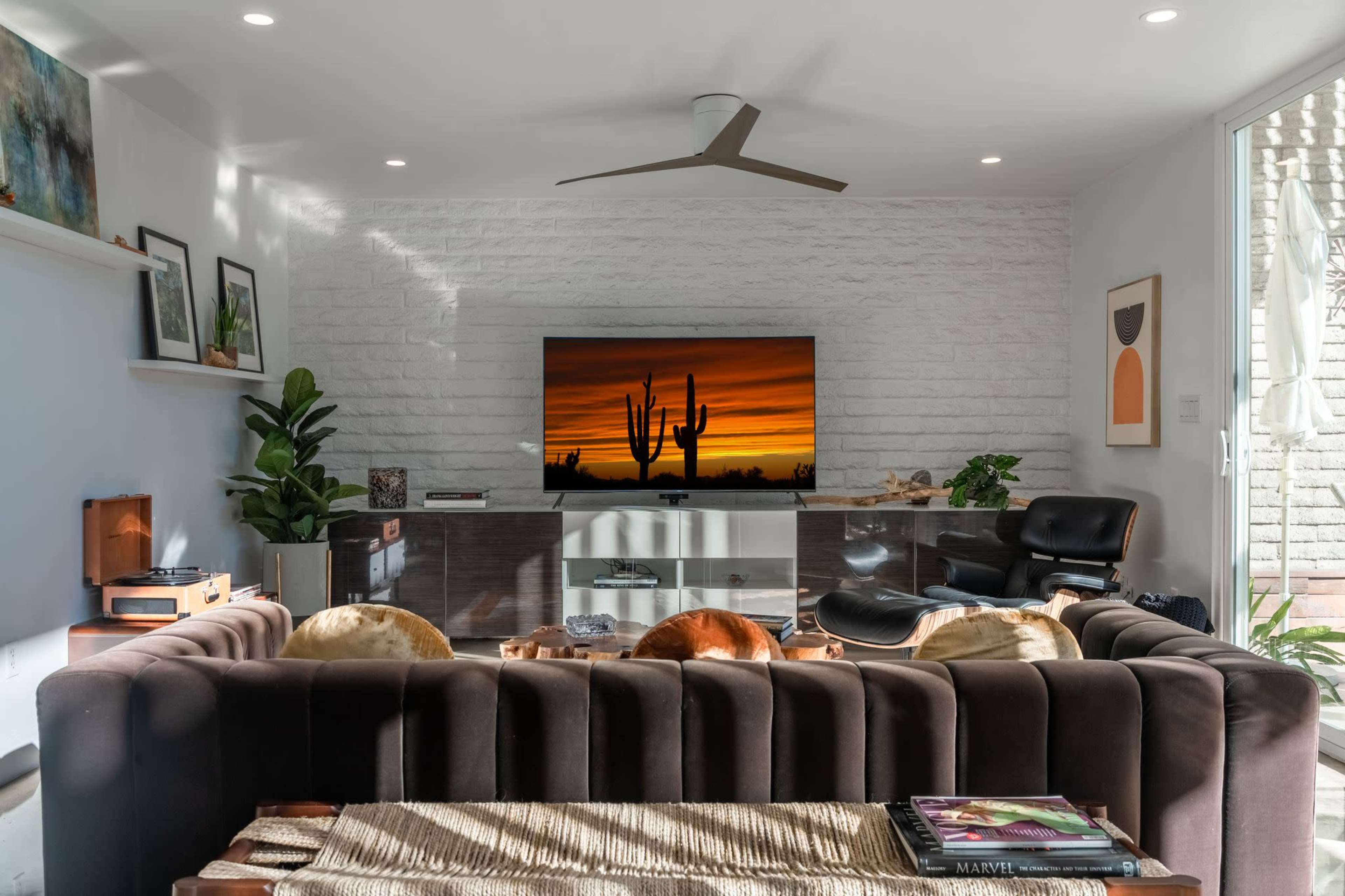 A modern living room features a dark sectional sofa facing a television displaying a sunset over cacti, surrounded by plants and minimalist decor.