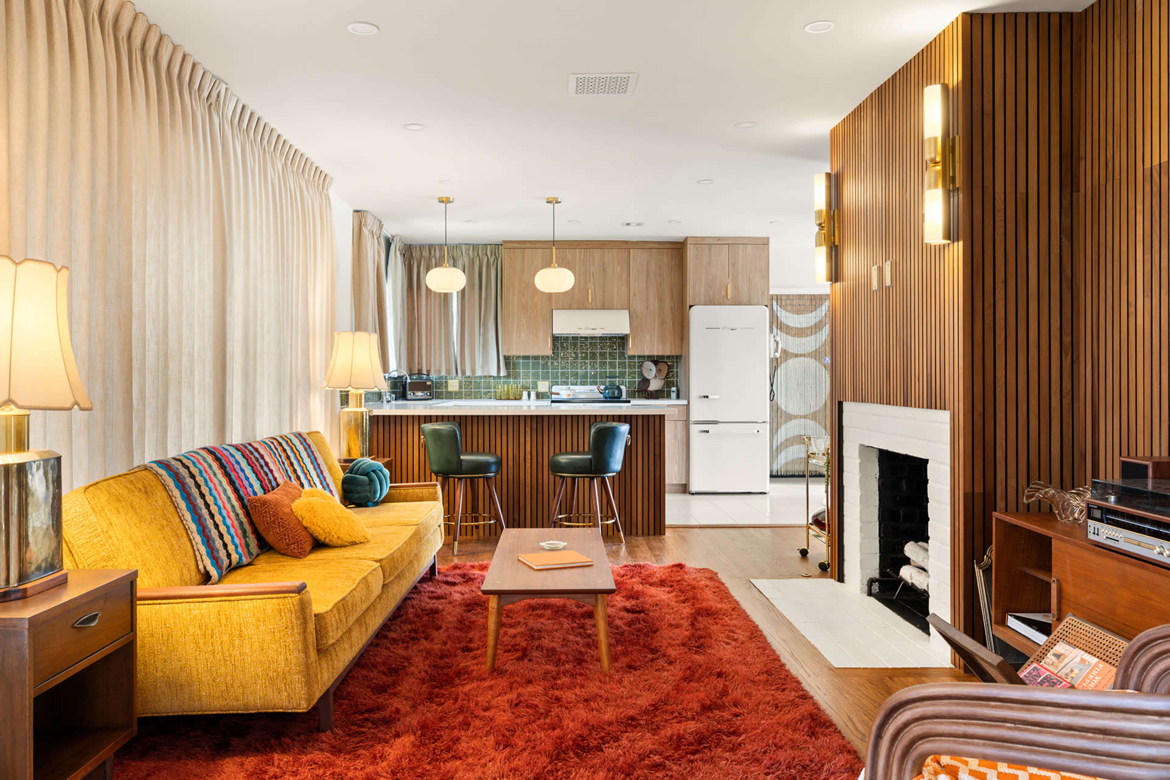 The image shows a modern living room with a yellow sofa, a wooden coffee table, and a kitchen area in the background featuring green tiles and white appliances.
