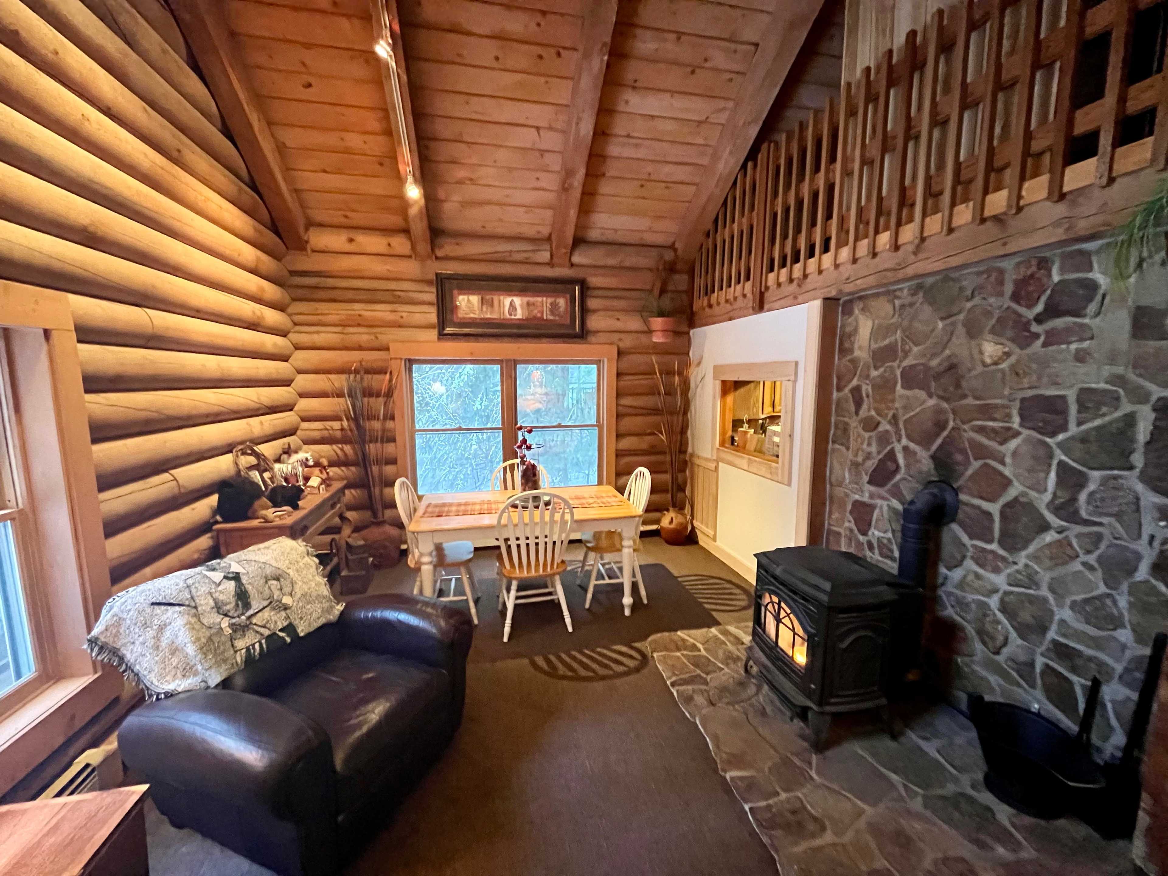 A rustic cabin interior with wooden log walls, a stone fireplace, a dining table with chairs, and a cozy seating area.