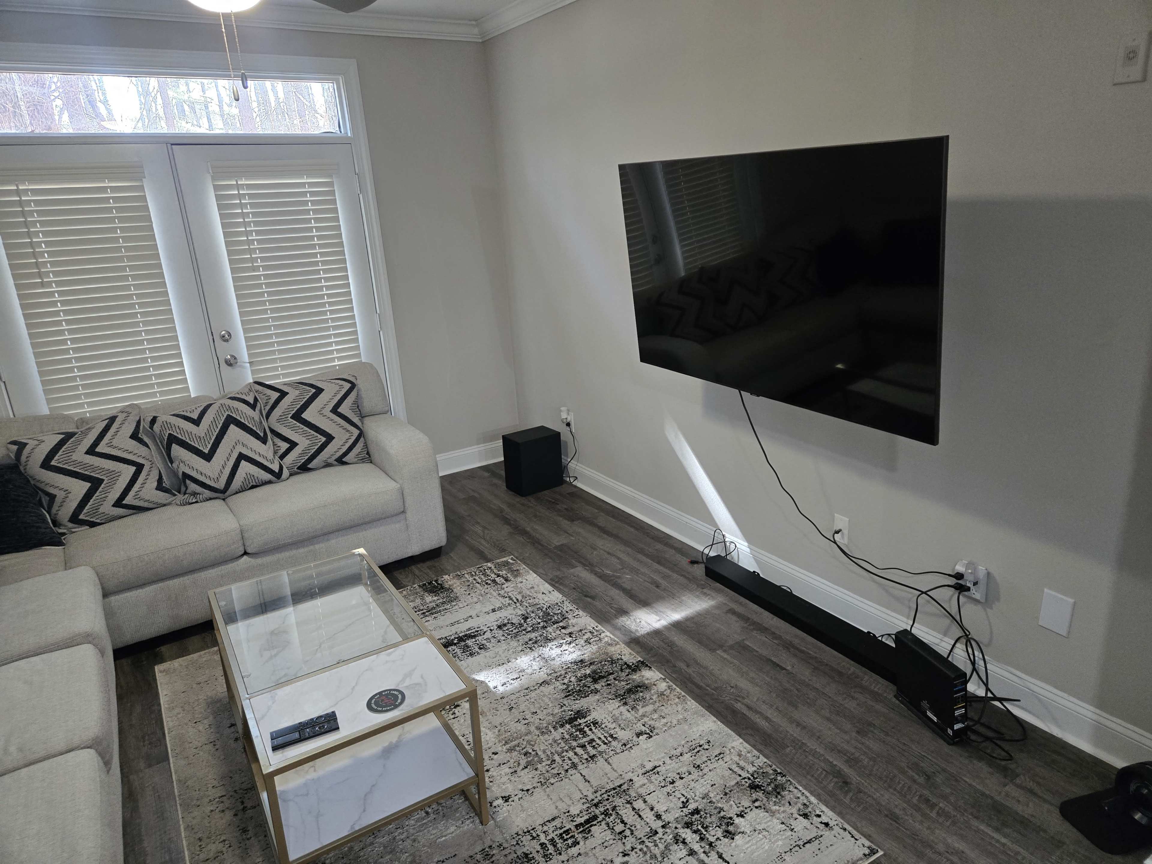 The image shows a modern living room with a gray sofa, a glass coffee table, and a mounted flat-screen TV on the wall.