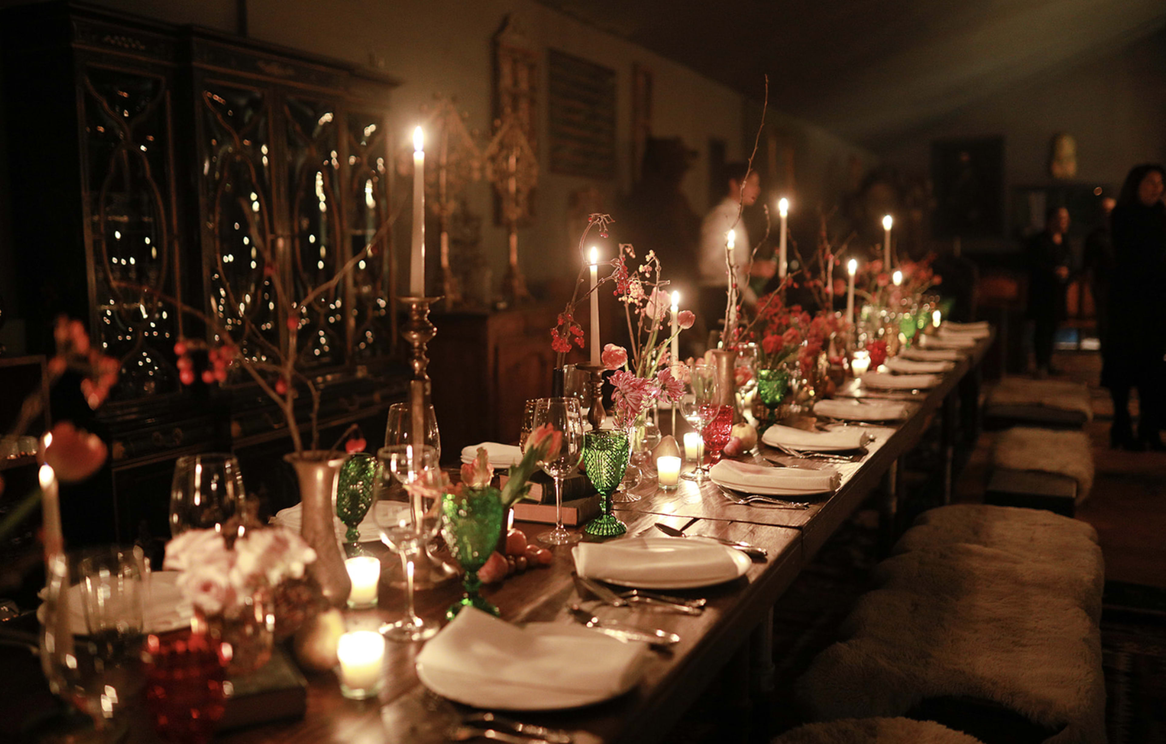 A long wooden dining table is elegantly set with candles, decorative flowers, and glassware in a dimly lit room.