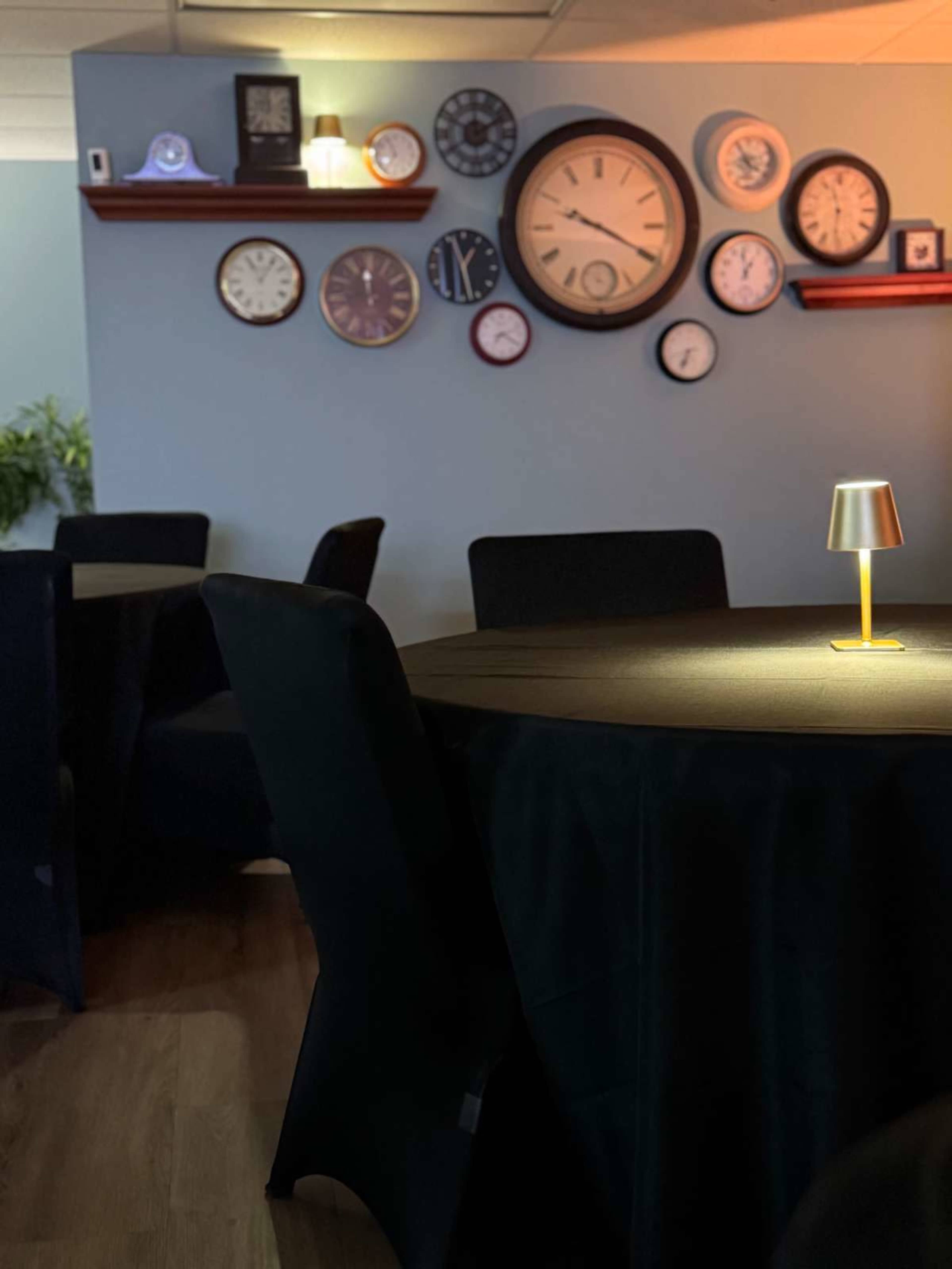 A dimly lit room features several clocks on the wall and empty tables covered with dark tablecloths, with a small lamp illuminating one table.