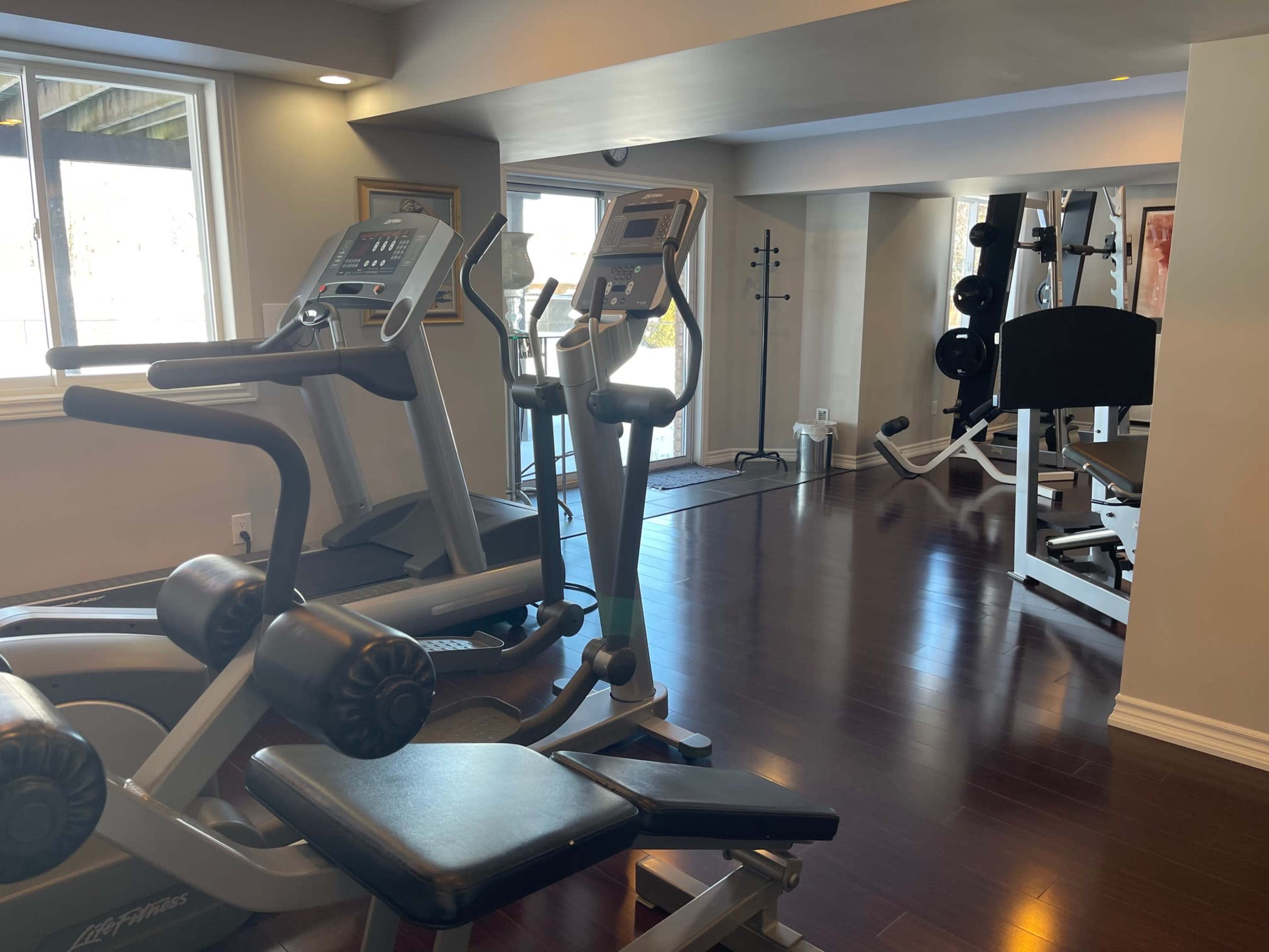 The image shows a bright home gym equipped with exercise machines, a weight bench, and large windows allowing natural light.