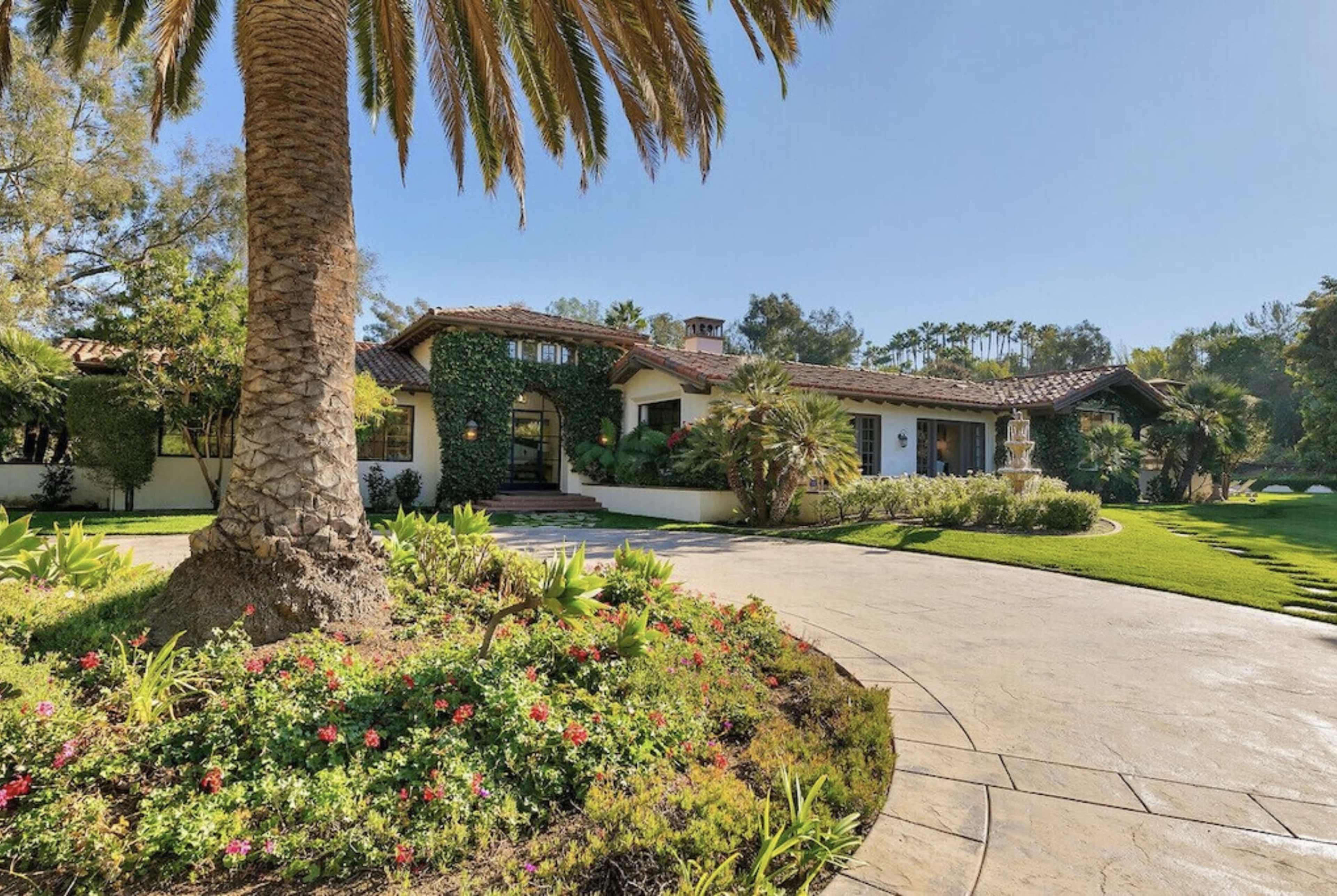 A Mediterranean-style home with a tile roof is surrounded by lush landscaping and palm trees.