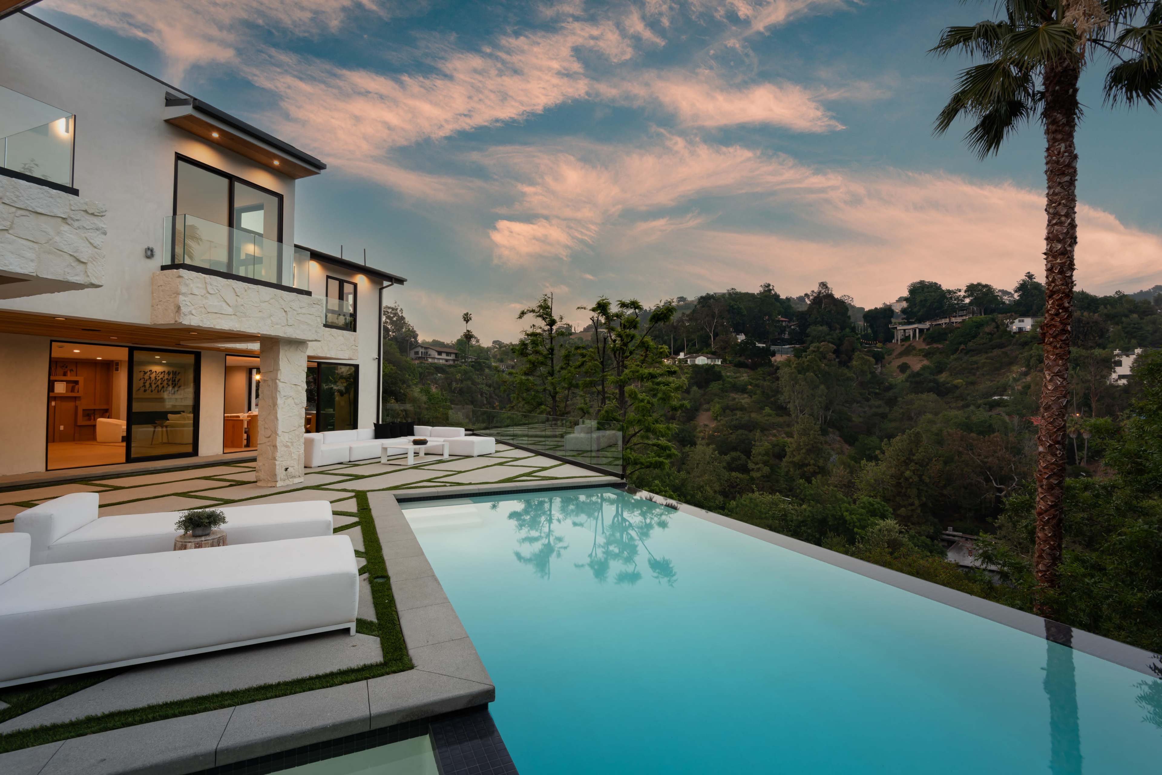 A modern villa with a sleek infinity pool overlooks a lush hillside at sunset.