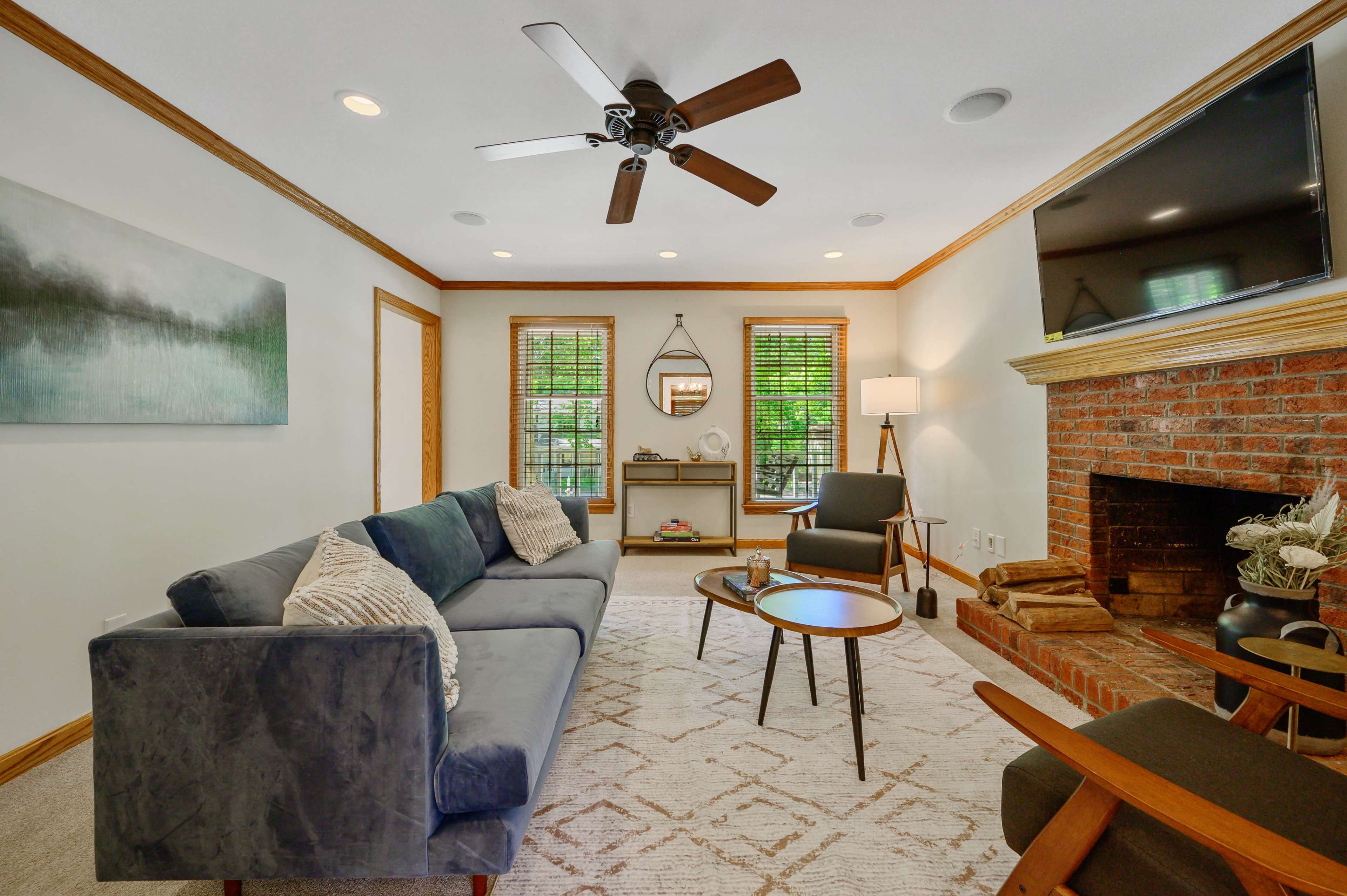 A cozy living room features a blue sofa, a brick fireplace, and large windows with greenery visible outside.