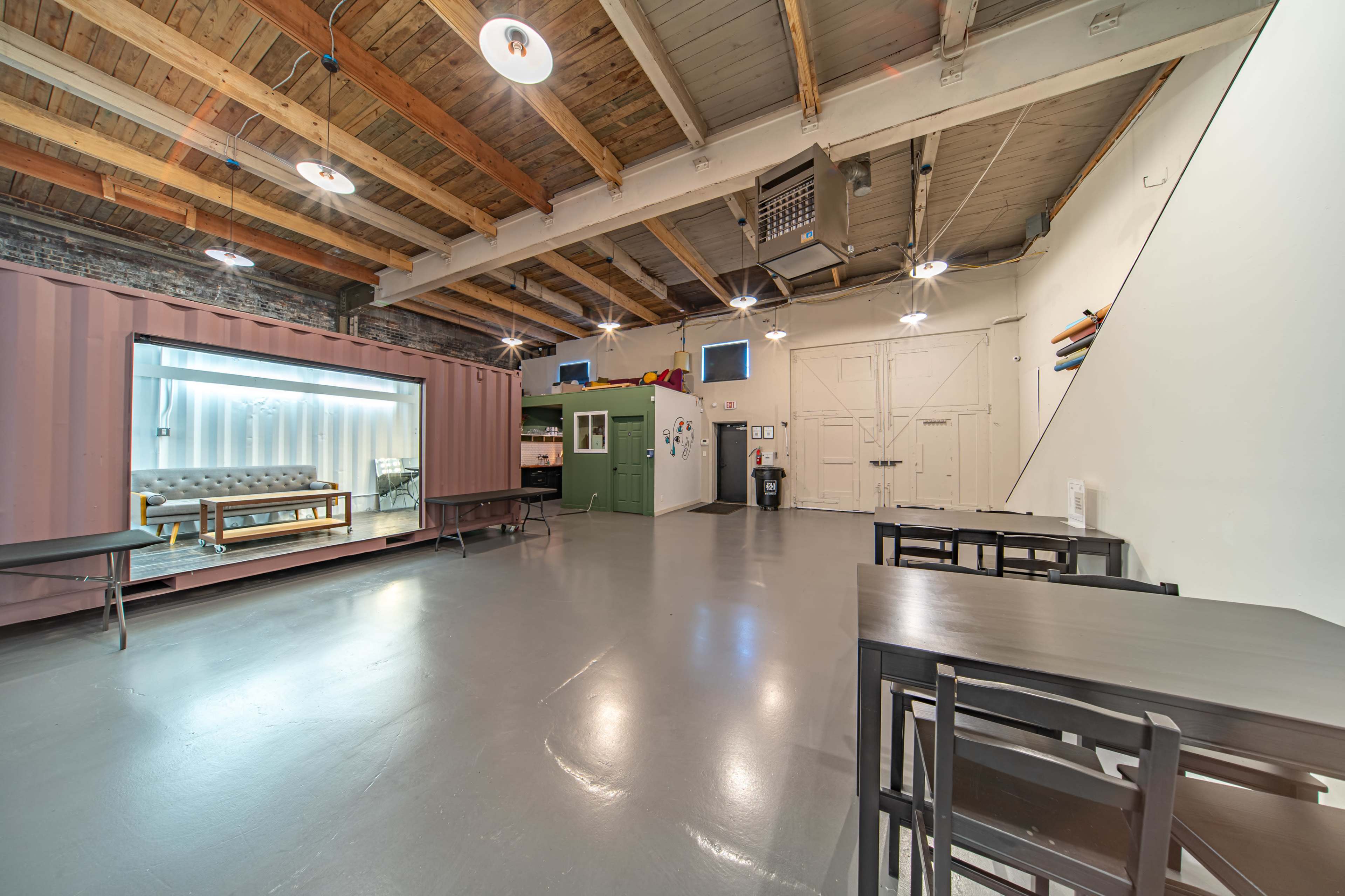 A spacious interior featuring exposed wooden beams, a shipping container, a green cabinet, and several tables arranged on a smooth, gray floor.