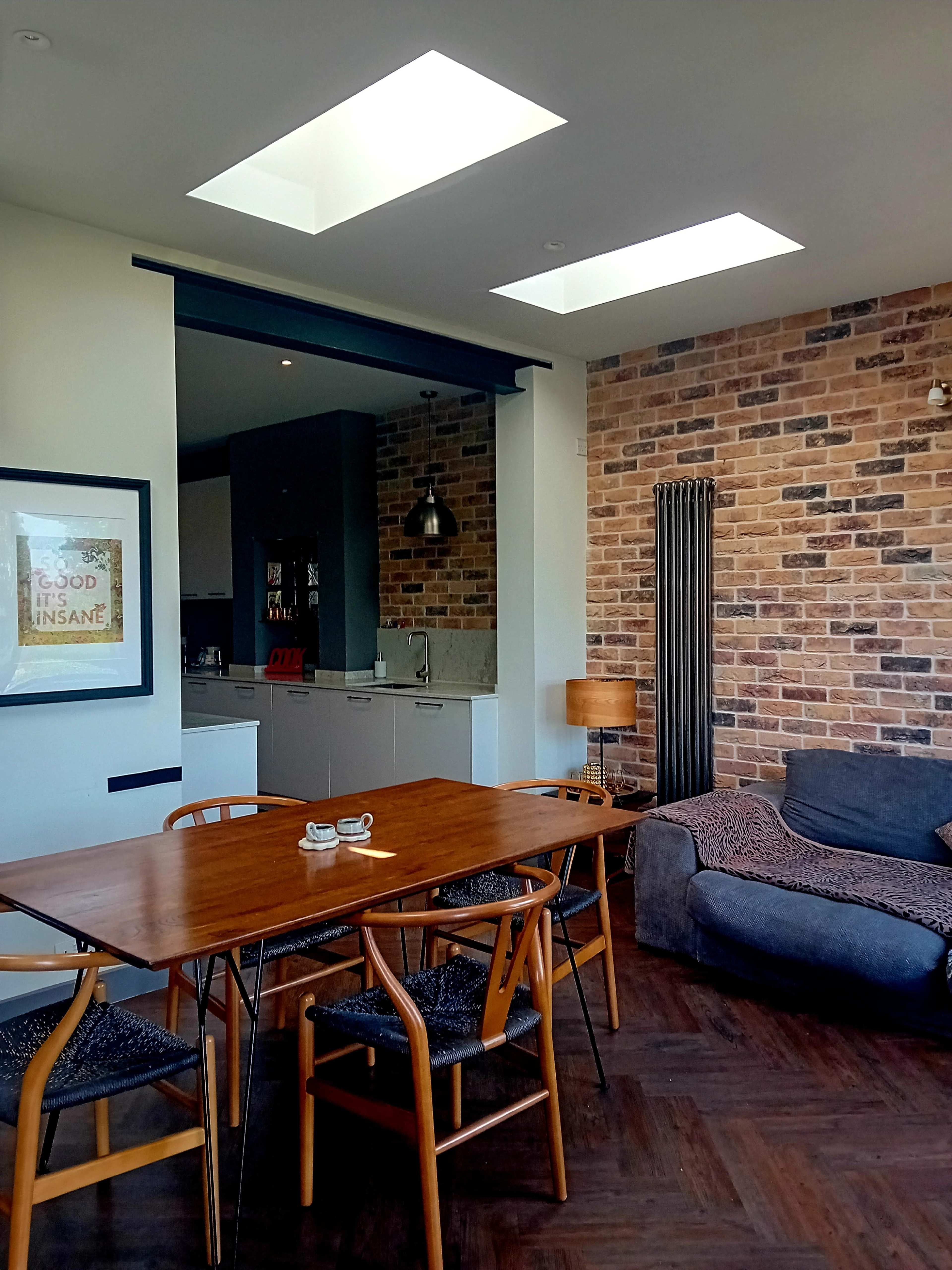 The image shows a modern living space with a wooden dining table, a gray sofa, and a kitchen area featuring a brick wall and skylights above.