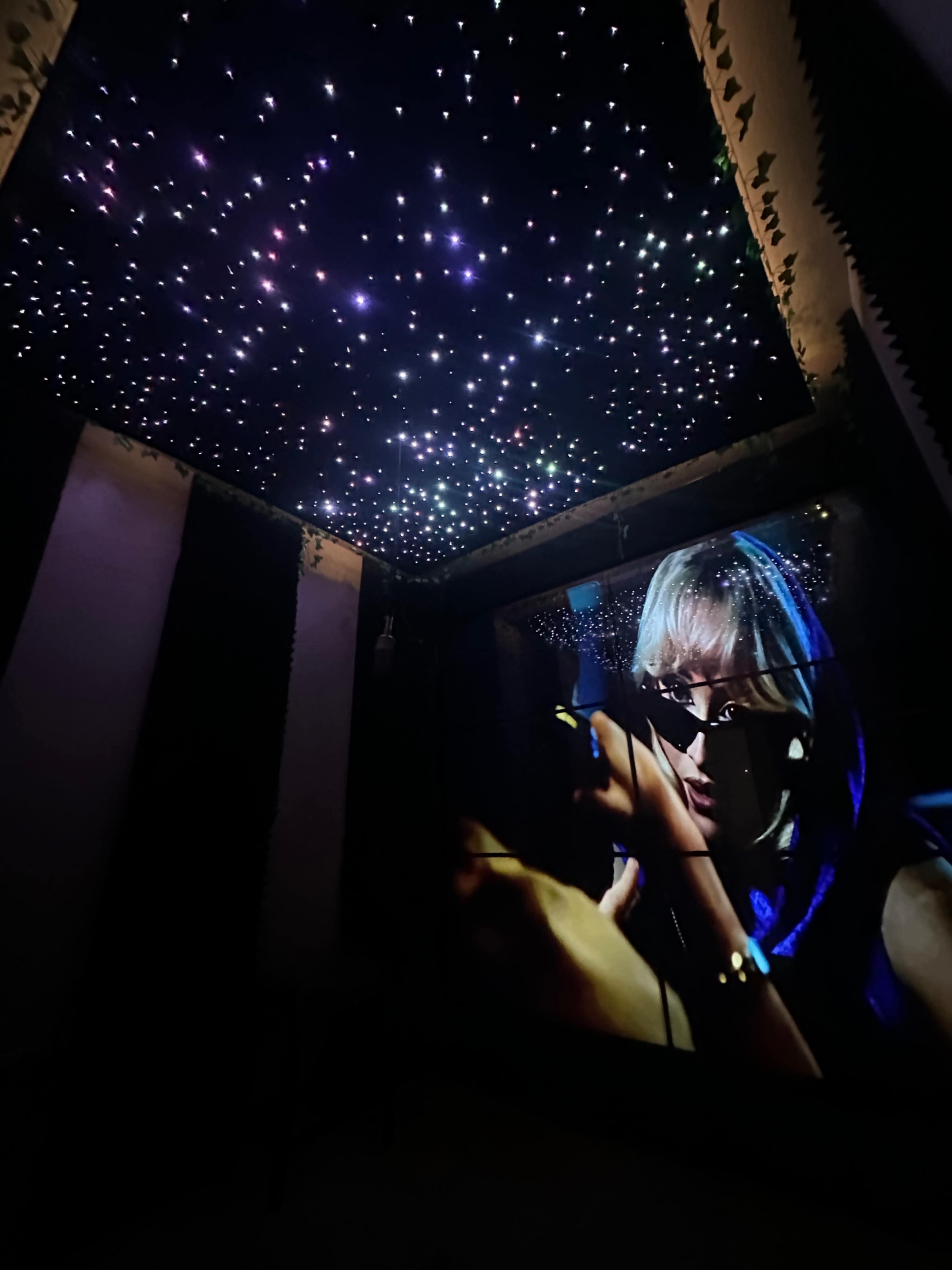 The image shows a dimly lit room with a starry ceiling projection and a large screen displaying a close-up of a person wearing sunglasses.
