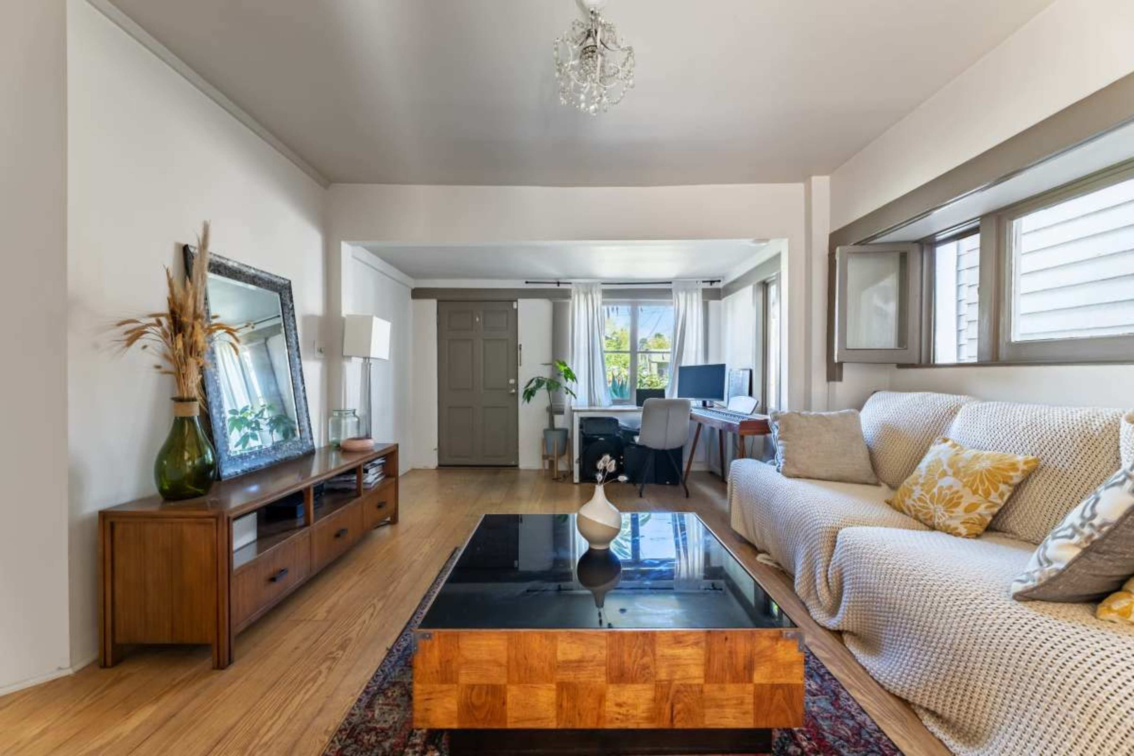 The room features a sofa, a wooden coffee table, a large mirror, and a workspace with a desk in a bright setting.