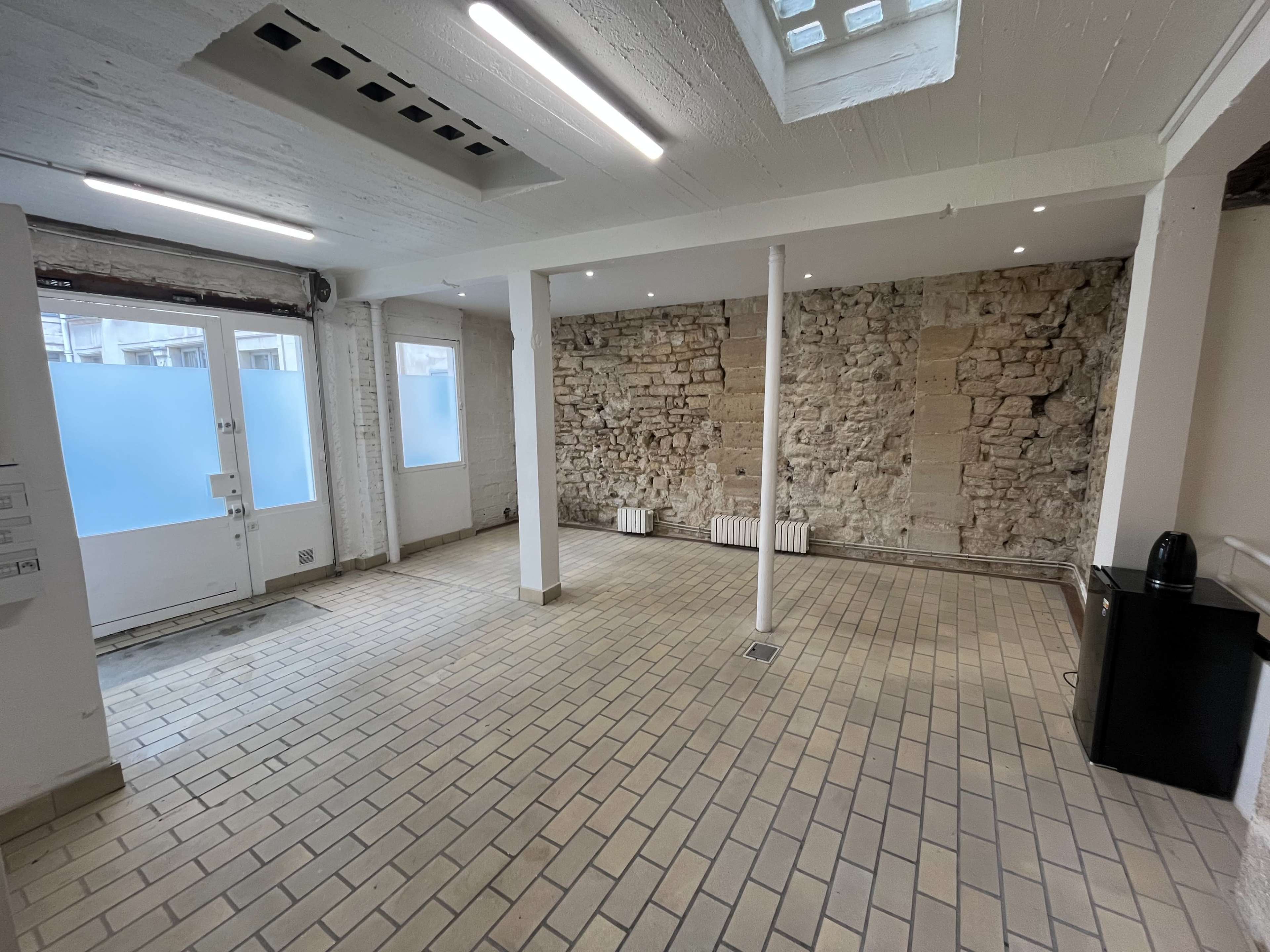The image shows a spacious room with stone walls, tiled flooring, and large windows allowing natural light.