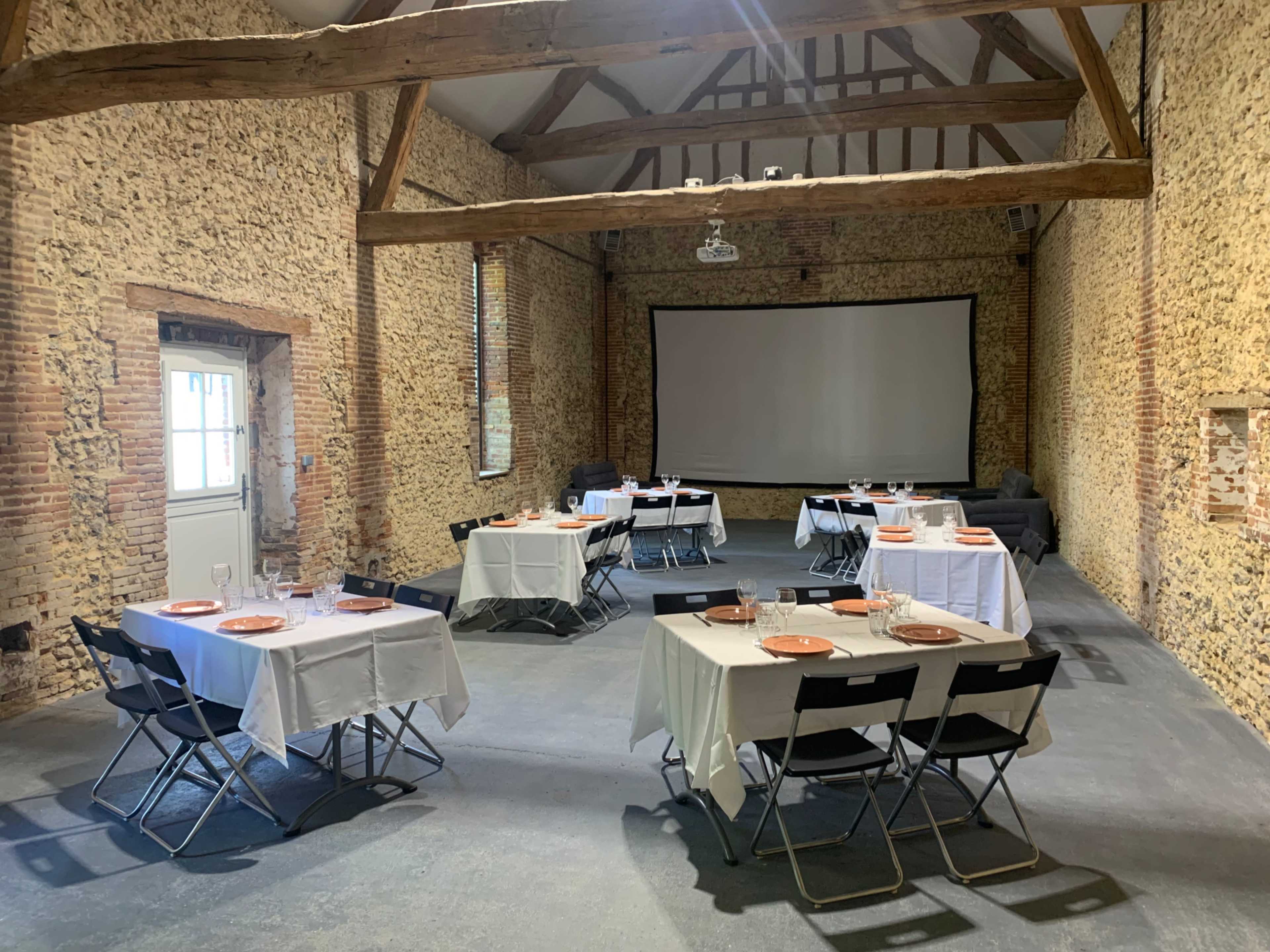 A spacious, rustic room with stone walls featuring several tables set for dining, each adorned with plates and glassware, and a projector screen at one end.