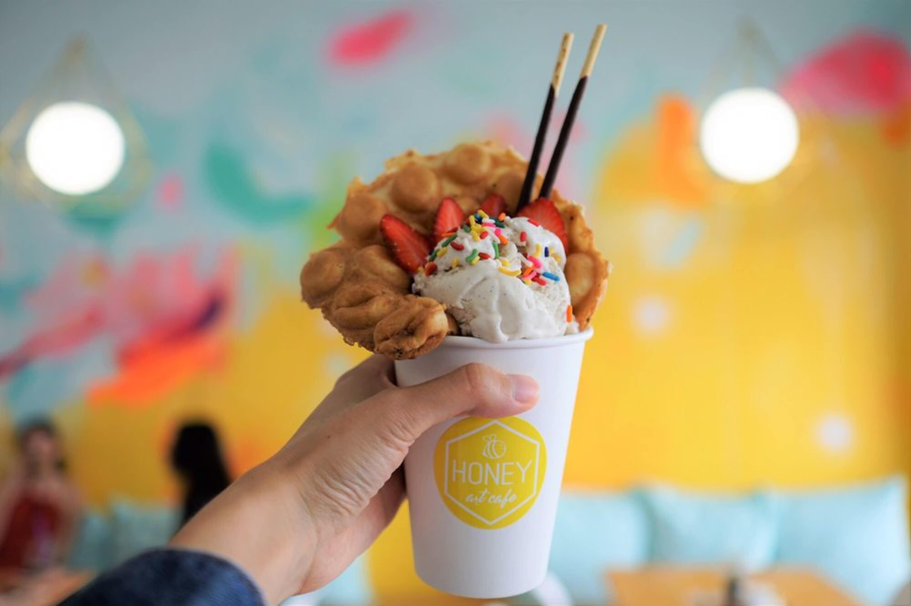 A hand holds a cup filled with ice cream topped with strawberries and sprinkles, with decorative chopsticks, against a colorful background.