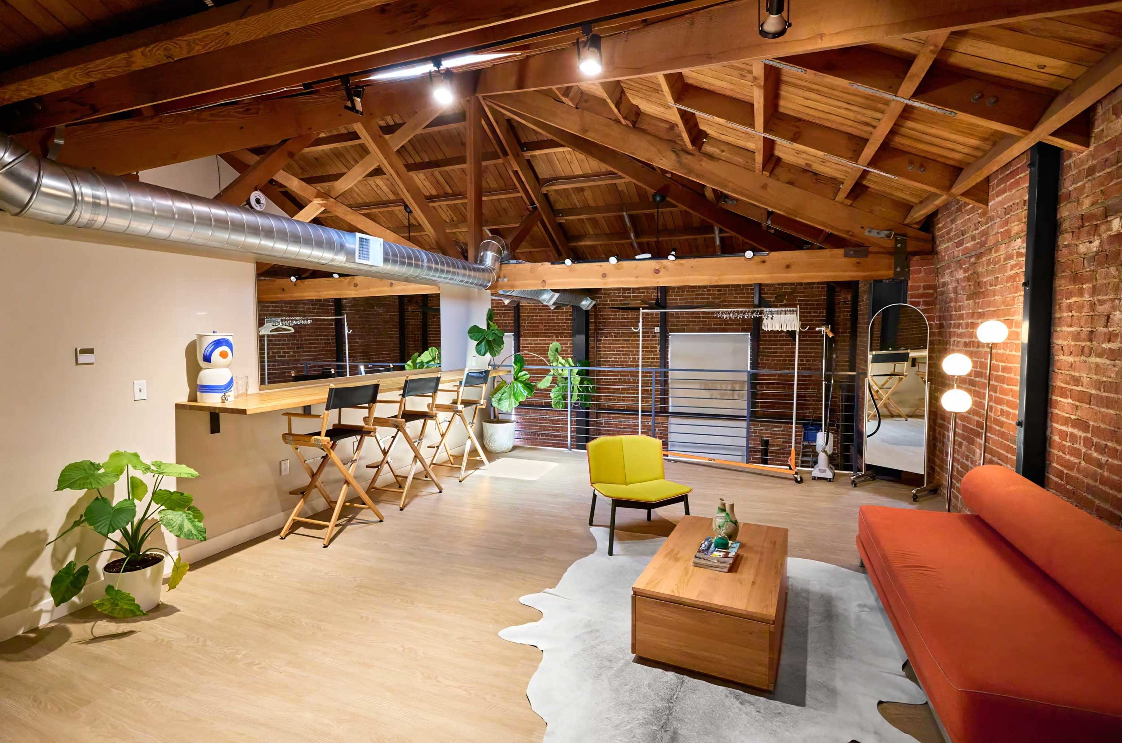 The image shows a modern loft-style living space with exposed brick walls, wooden beams, a bar with stools, and a yellow armchair.