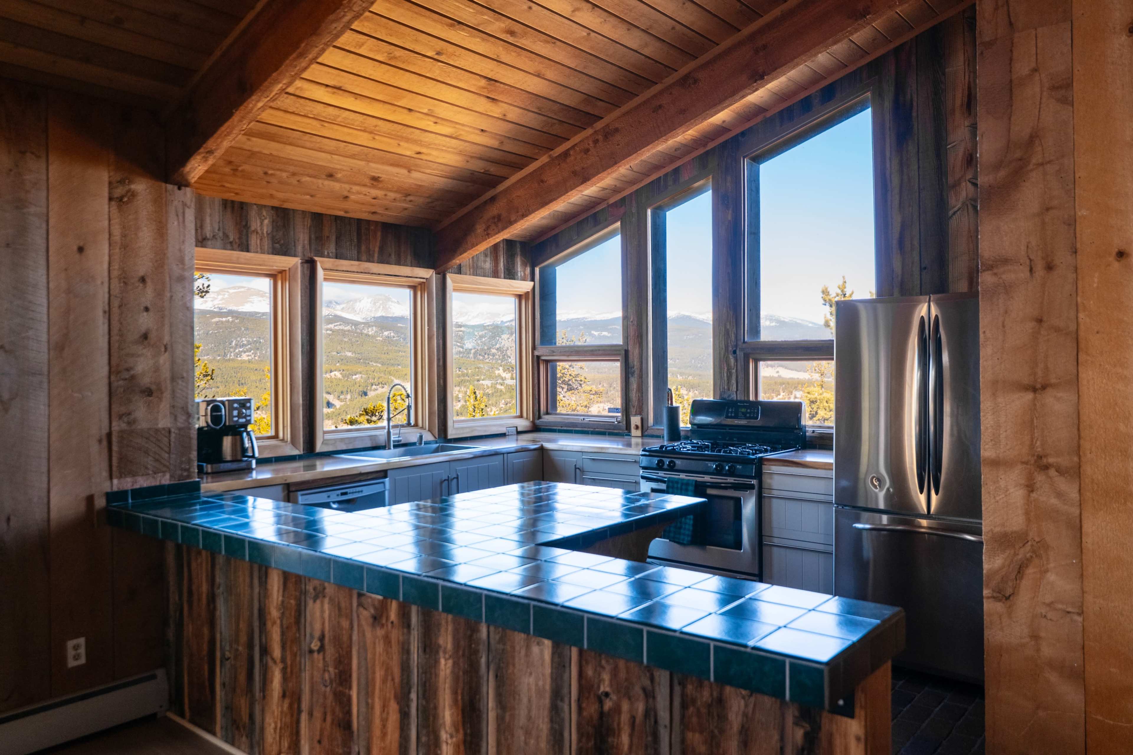 The kitchen features a wooden ceiling, large windows with mountain views, and a tile-covered island and countertop.