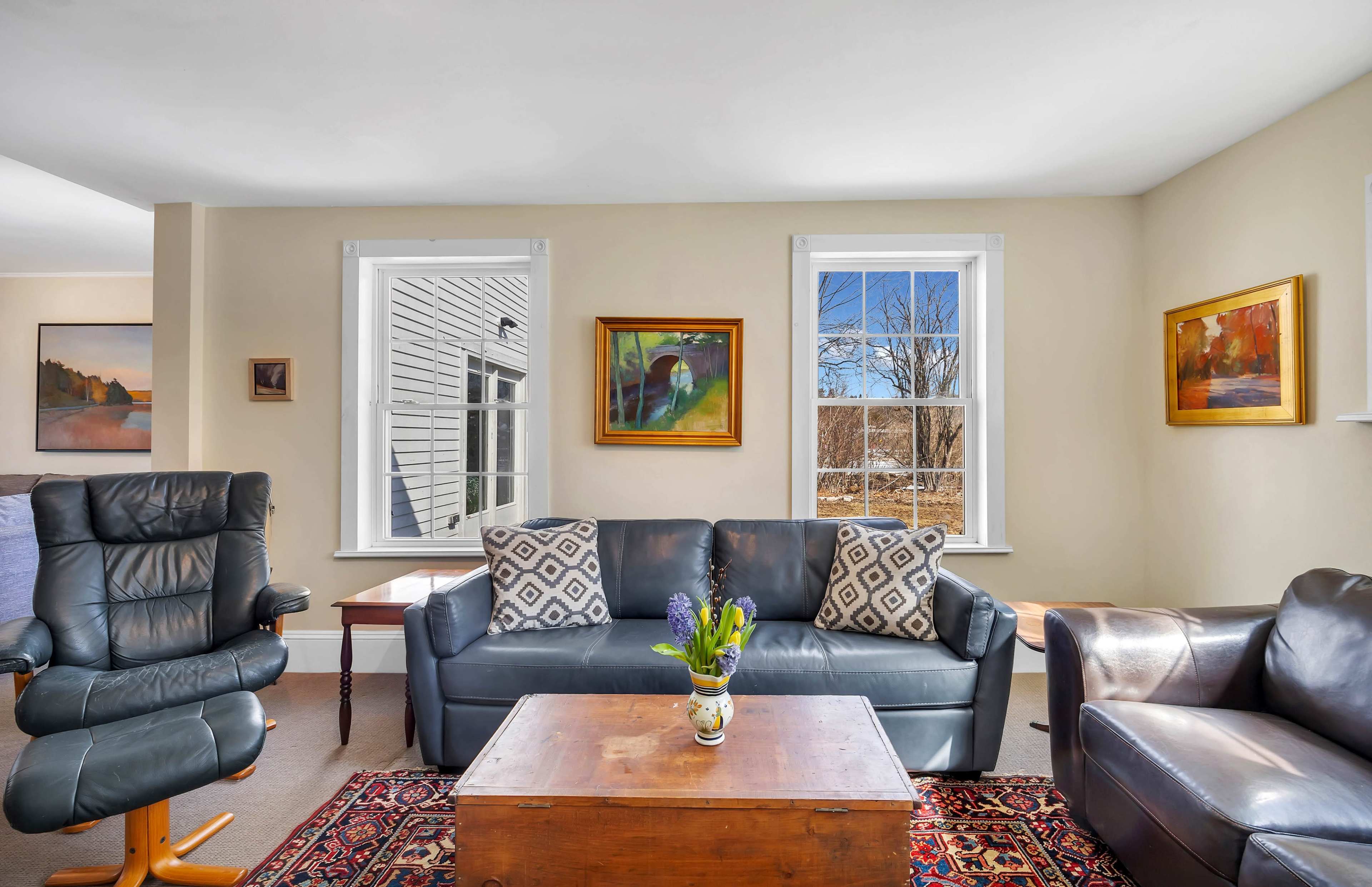 A cozy living room features a black leather couch and armchair, a wooden coffee table, and large windows that provide natural light.