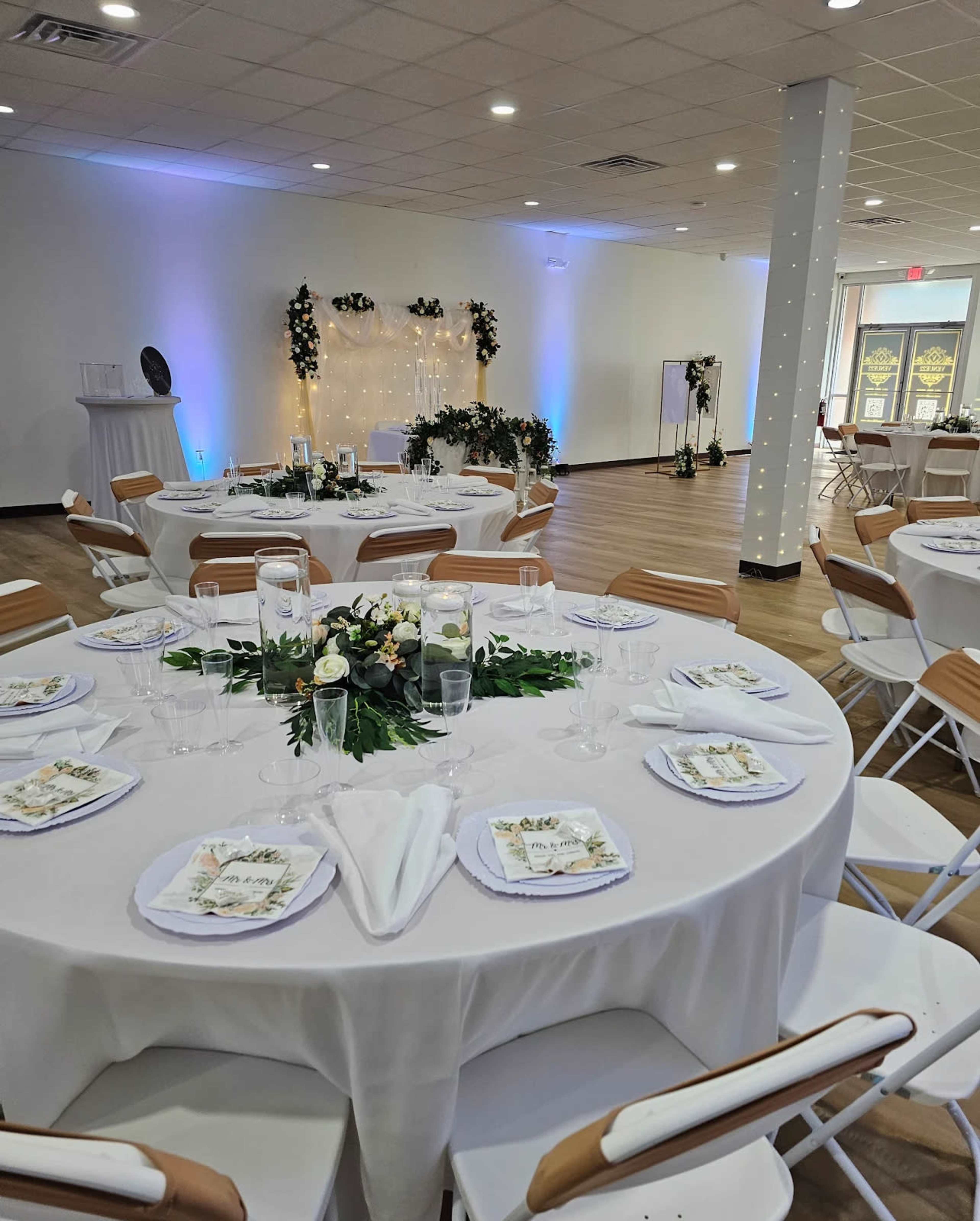 The image shows a banquet hall set up for an event, featuring round tables with white tablecloths, floral centerpieces, and a decorated backdrop illuminated with soft lighting.