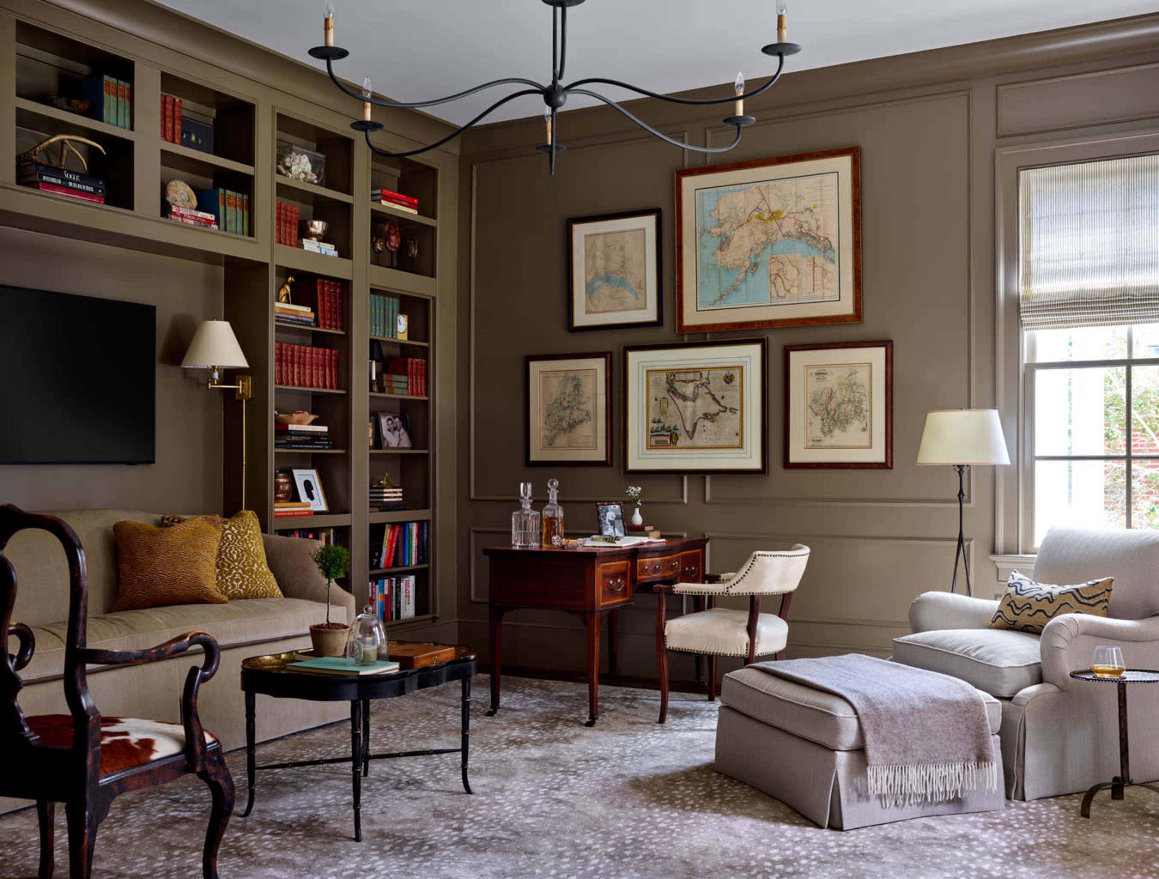 A cozy living room with a dark wall, a bookshelf filled with books, framed maps on display, and a mix of classic and modern furniture.