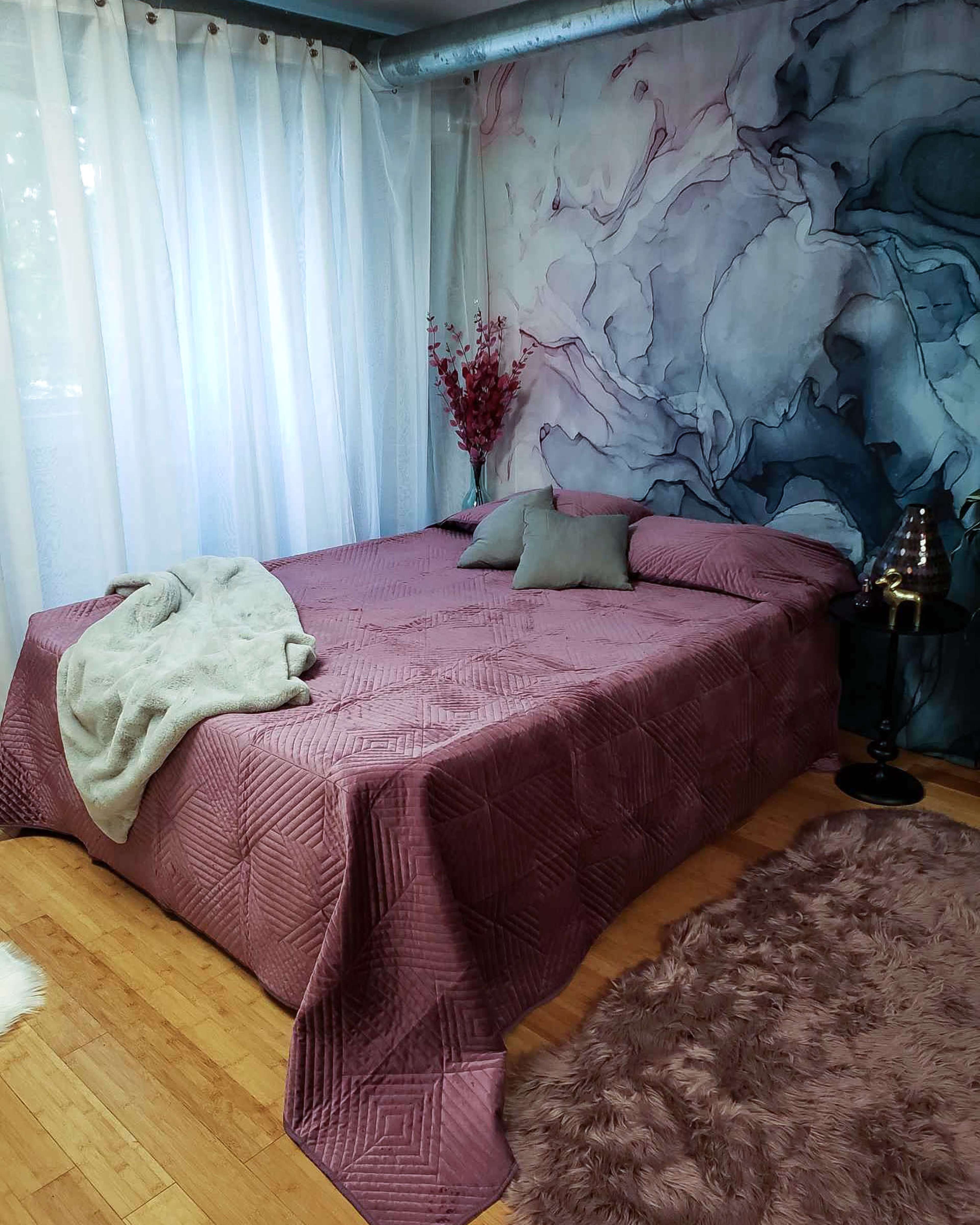 A neatly made bed with a mauve blanket and pillows is positioned against a wall featuring a flower-patterned mural, accompanied by sheer curtains and decorative items.