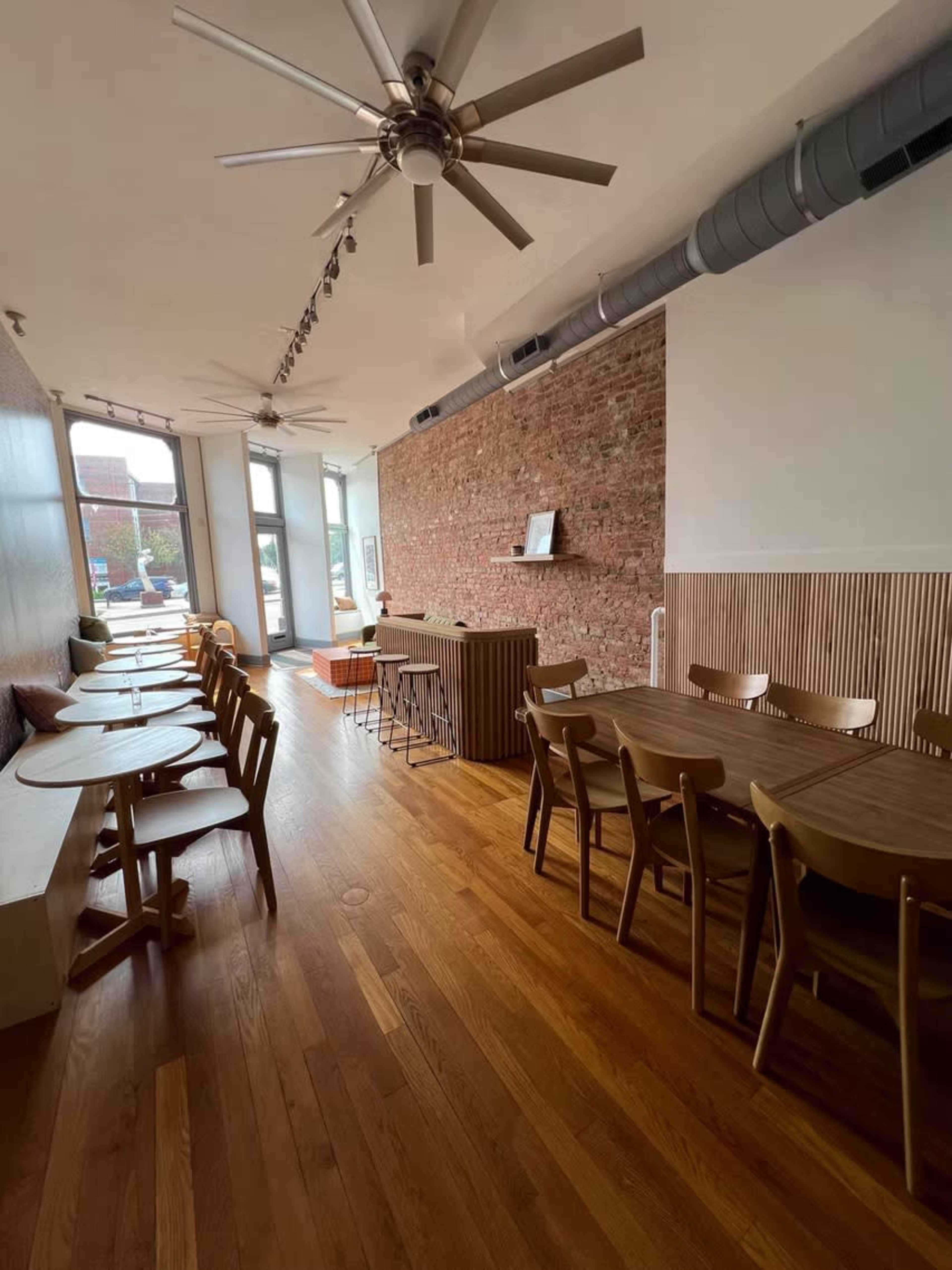The image shows a modern café interior with wooden flooring, a combination of seated tables and a service area, and a brick wall backdrop.