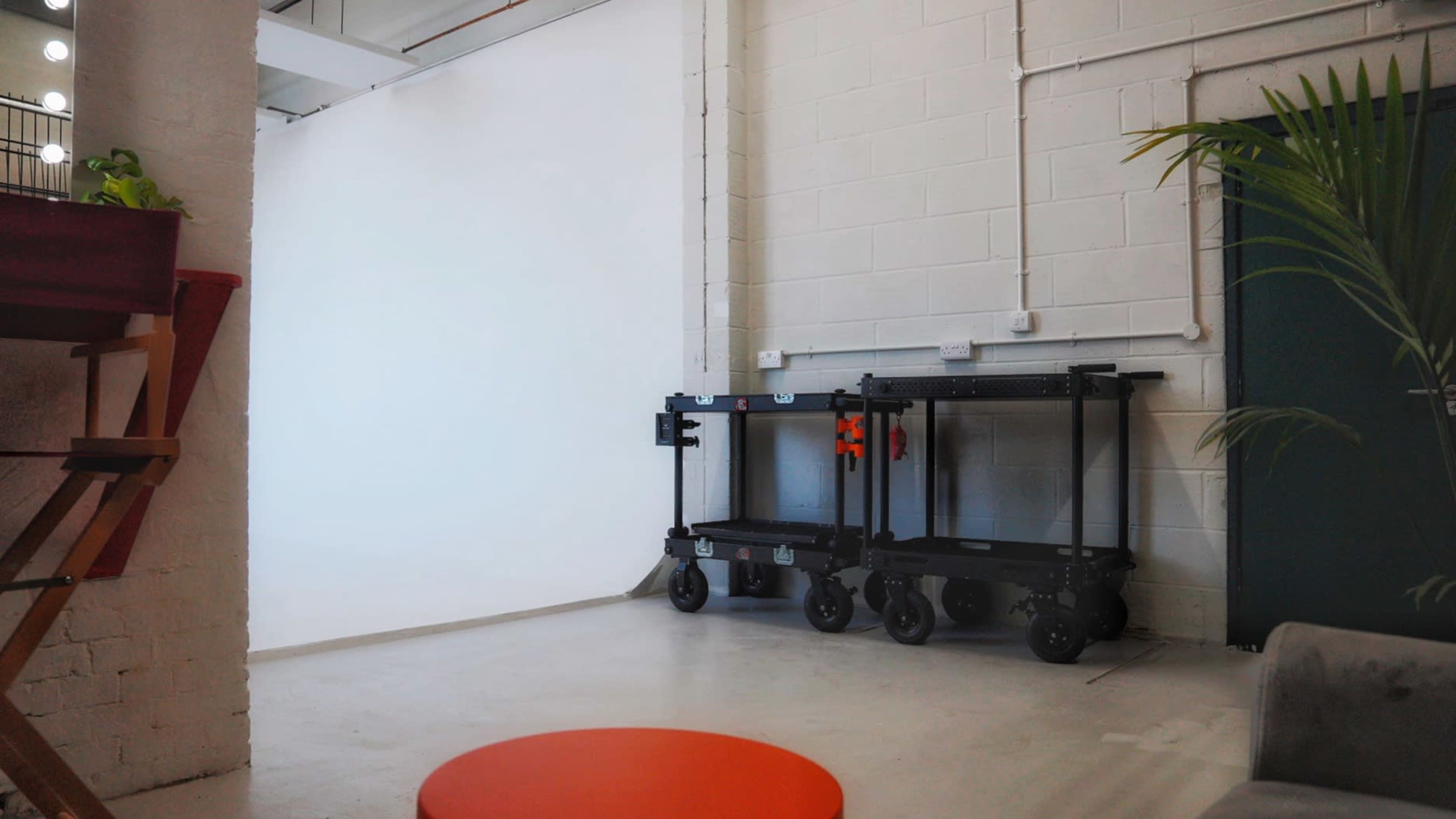 The image shows a spacious room with a white backdrop, two mobile carts against the wall, and a small orange table in the foreground.