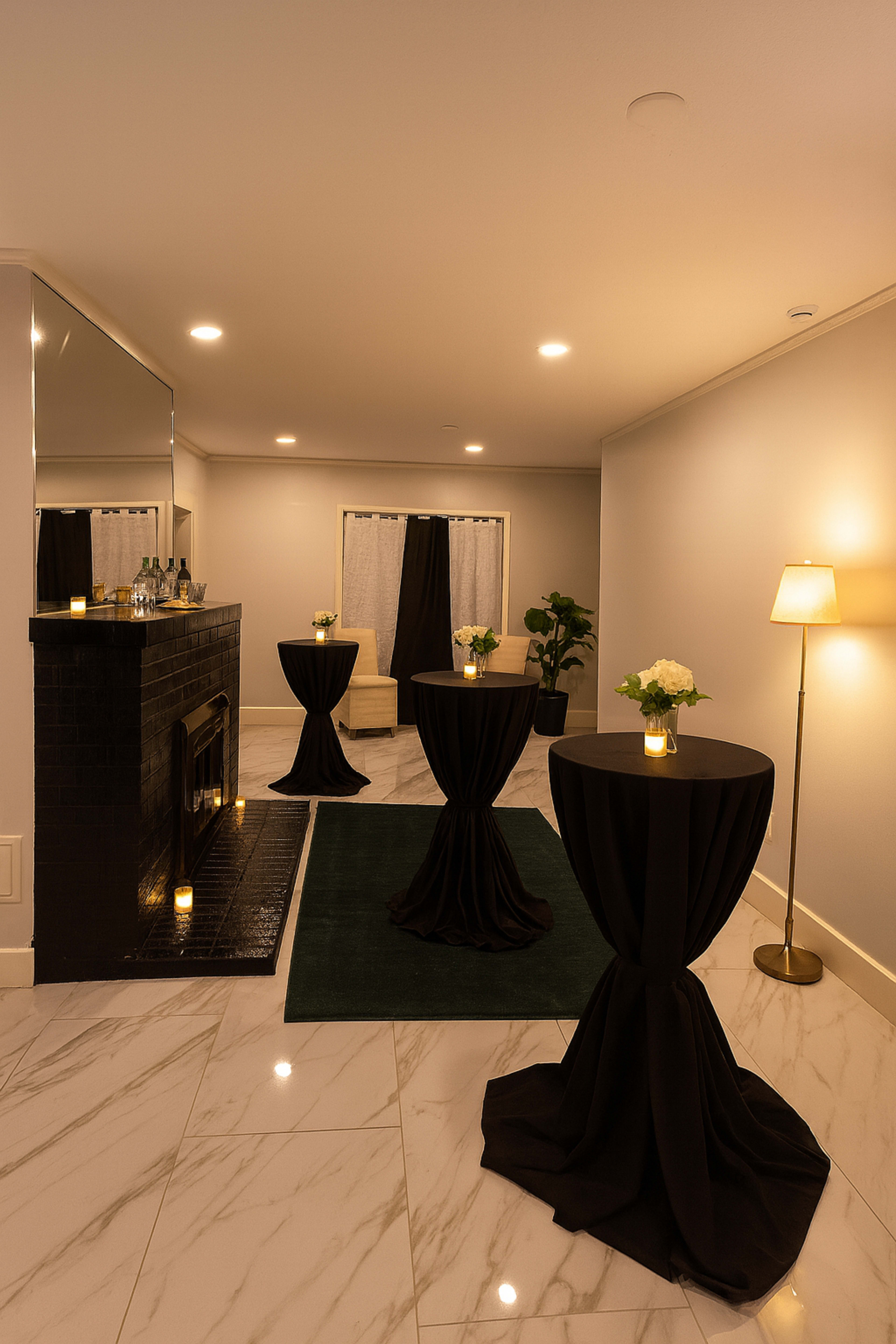 The image shows a stylish room featuring three tall, black-topped tables with floral centerpieces, a mirror, a fireplace, and soft lighting.