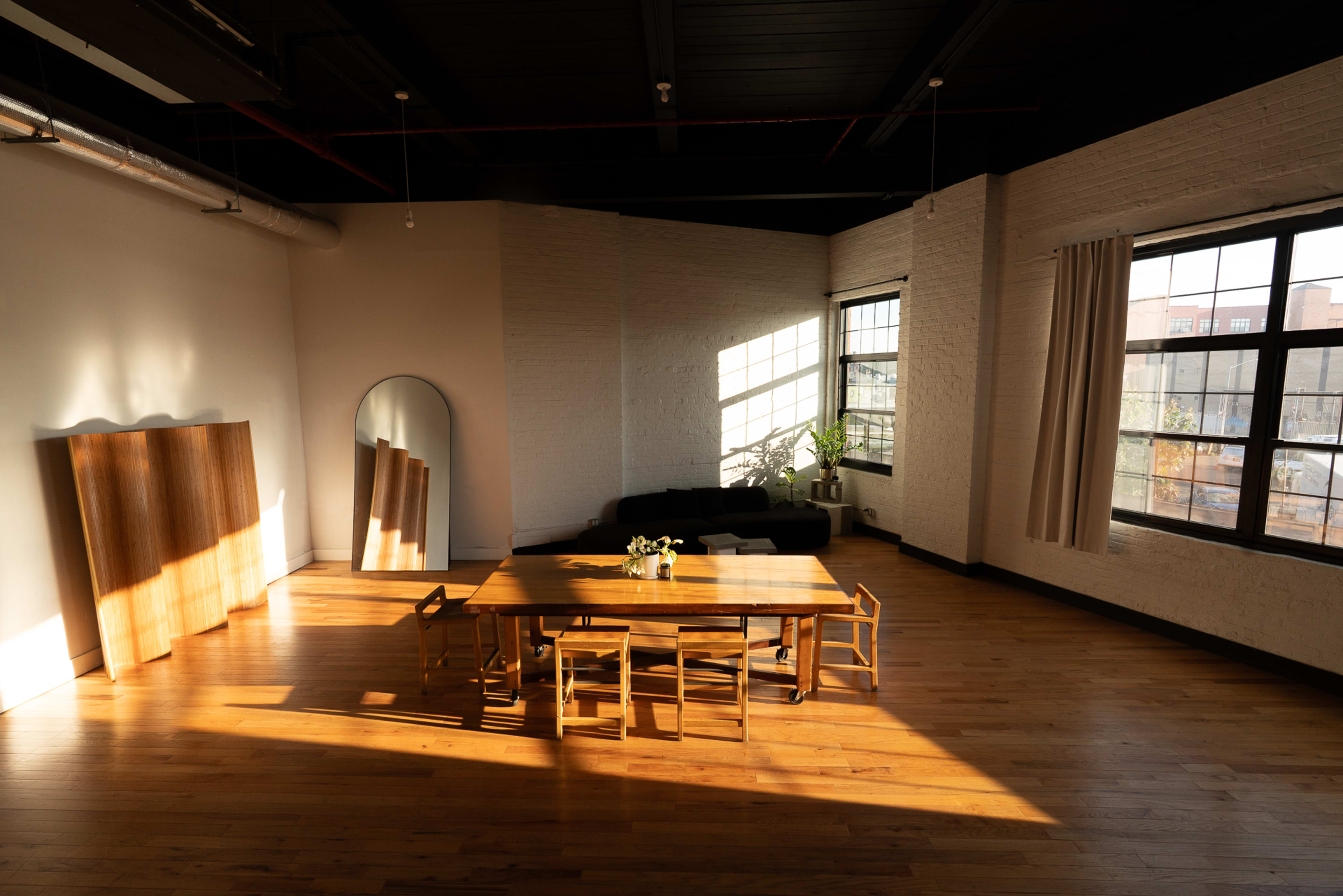 The image shows a spacious dining area with a wooden table and chairs, large windows allowing natural light, and a plant in the corner.