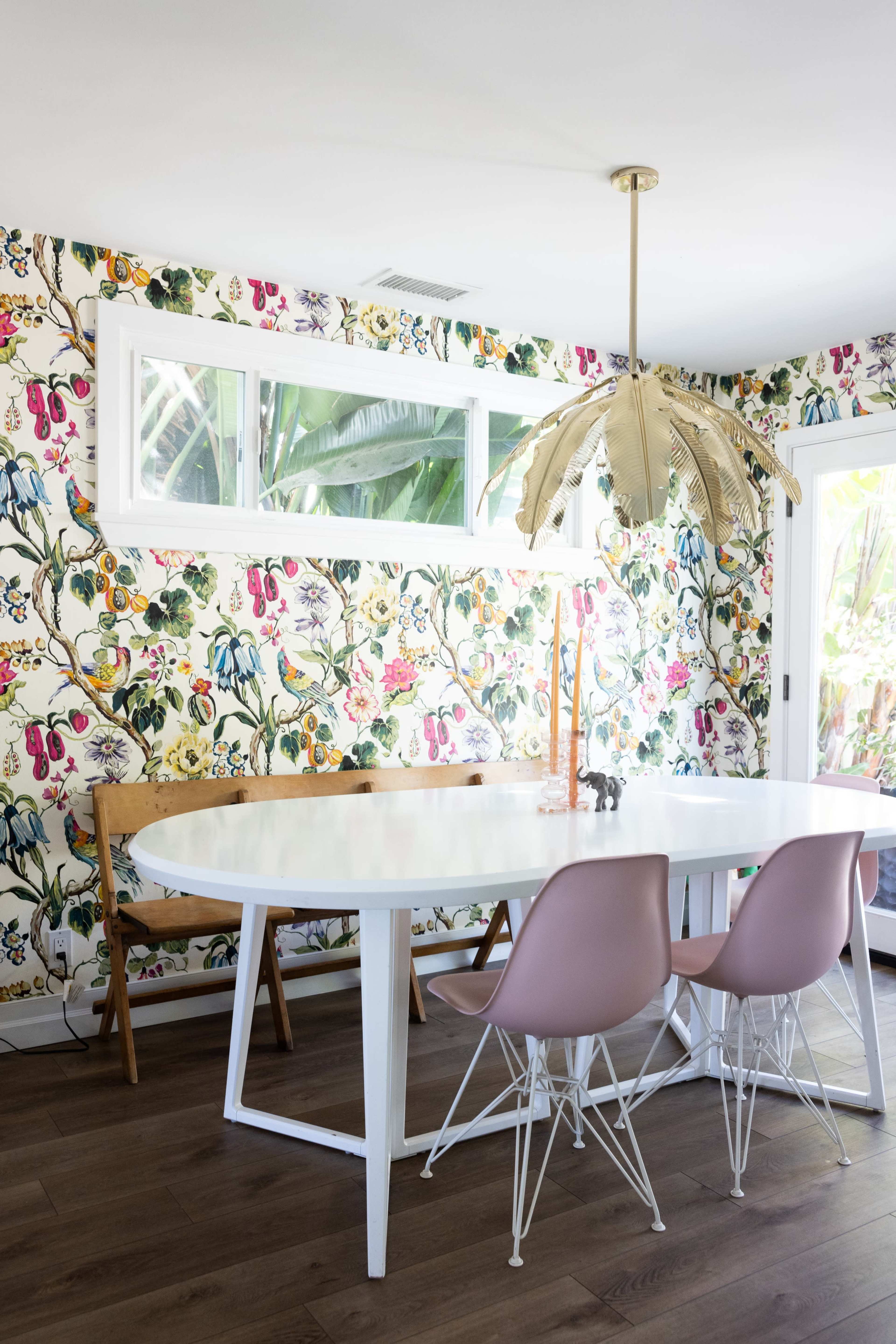 A modern dining area features a white oval table surrounded by pink chairs, set against a floral patterned wallpaper with a golden pendant light overhead.