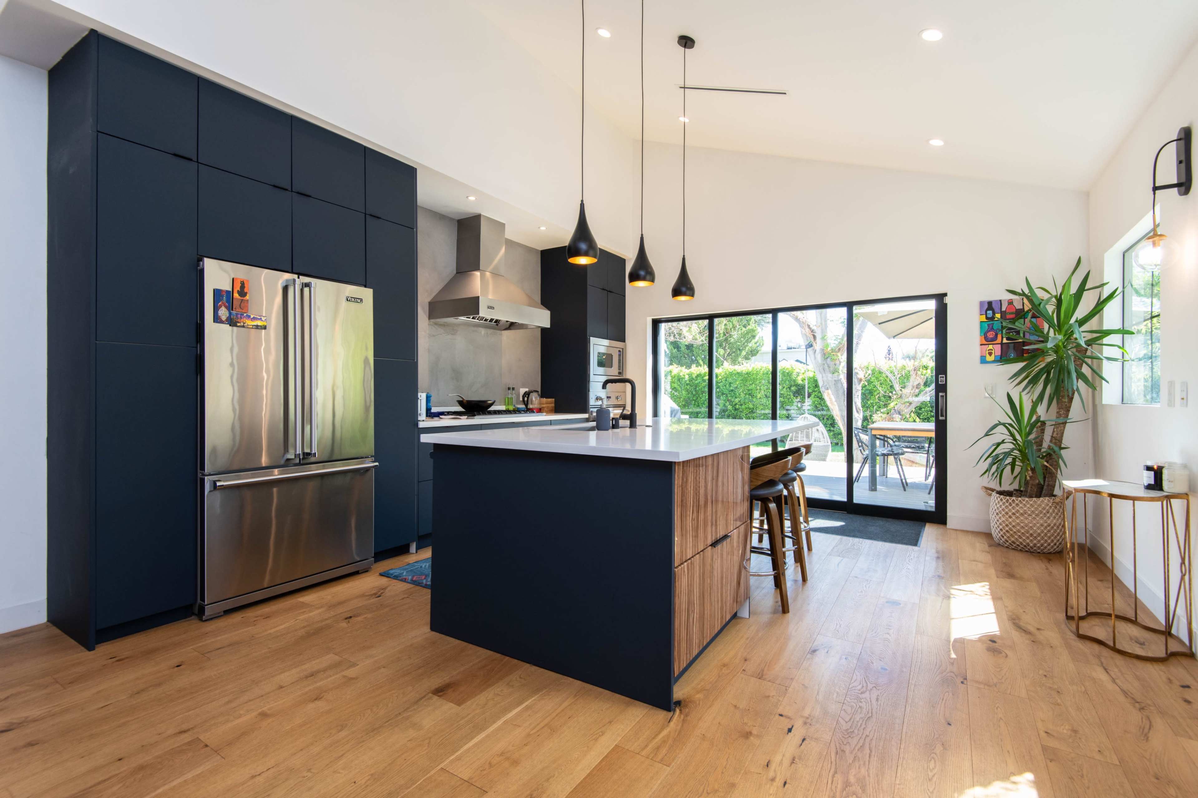 A modern kitchen with dark cabinets, a stainless steel refrigerator, and an island with wooden accents, leading to a patio area through large sliding glass doors.