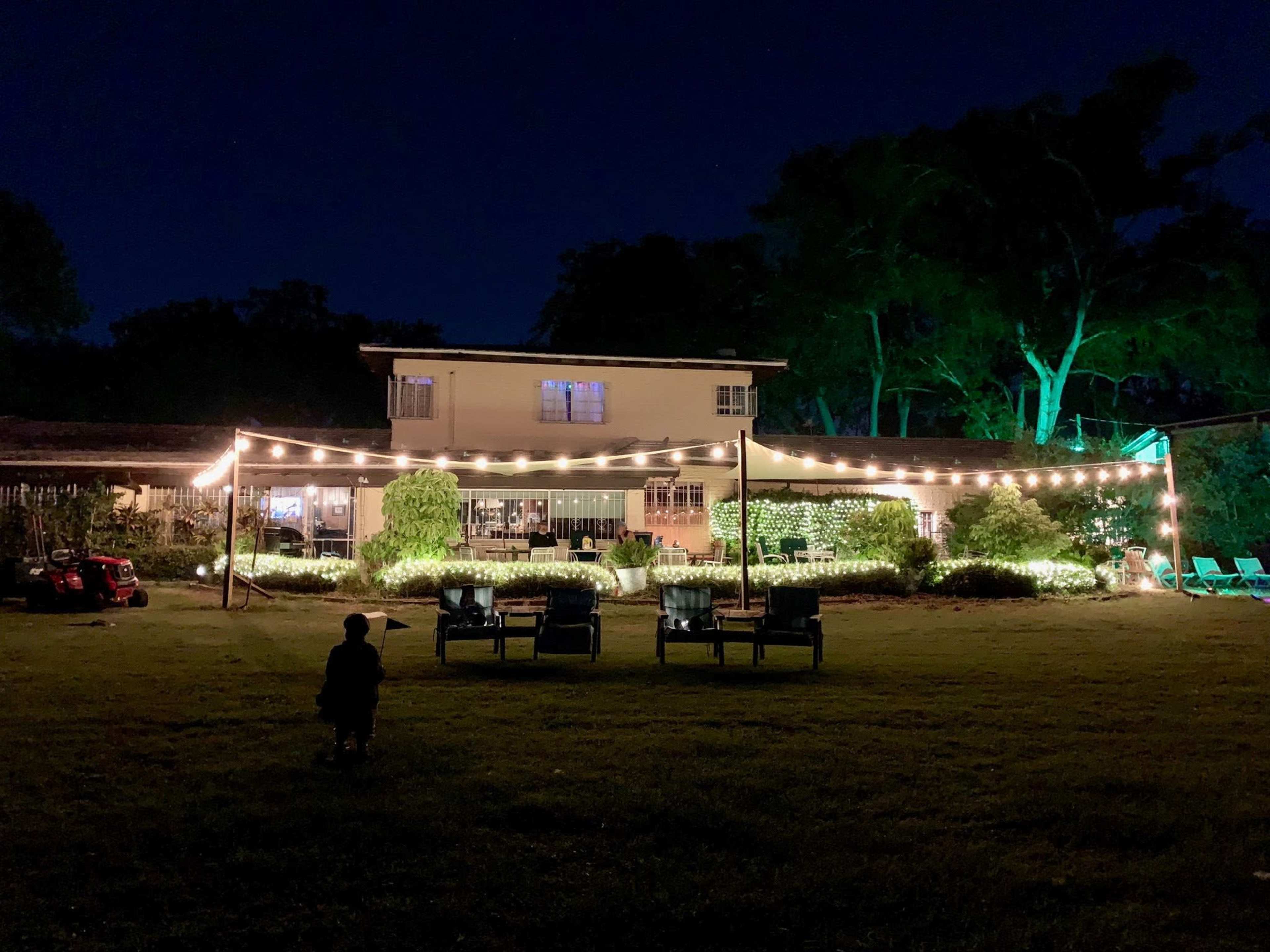A house is illuminated by string lights in a yard at night, with chairs arranged in front and trees in the background.