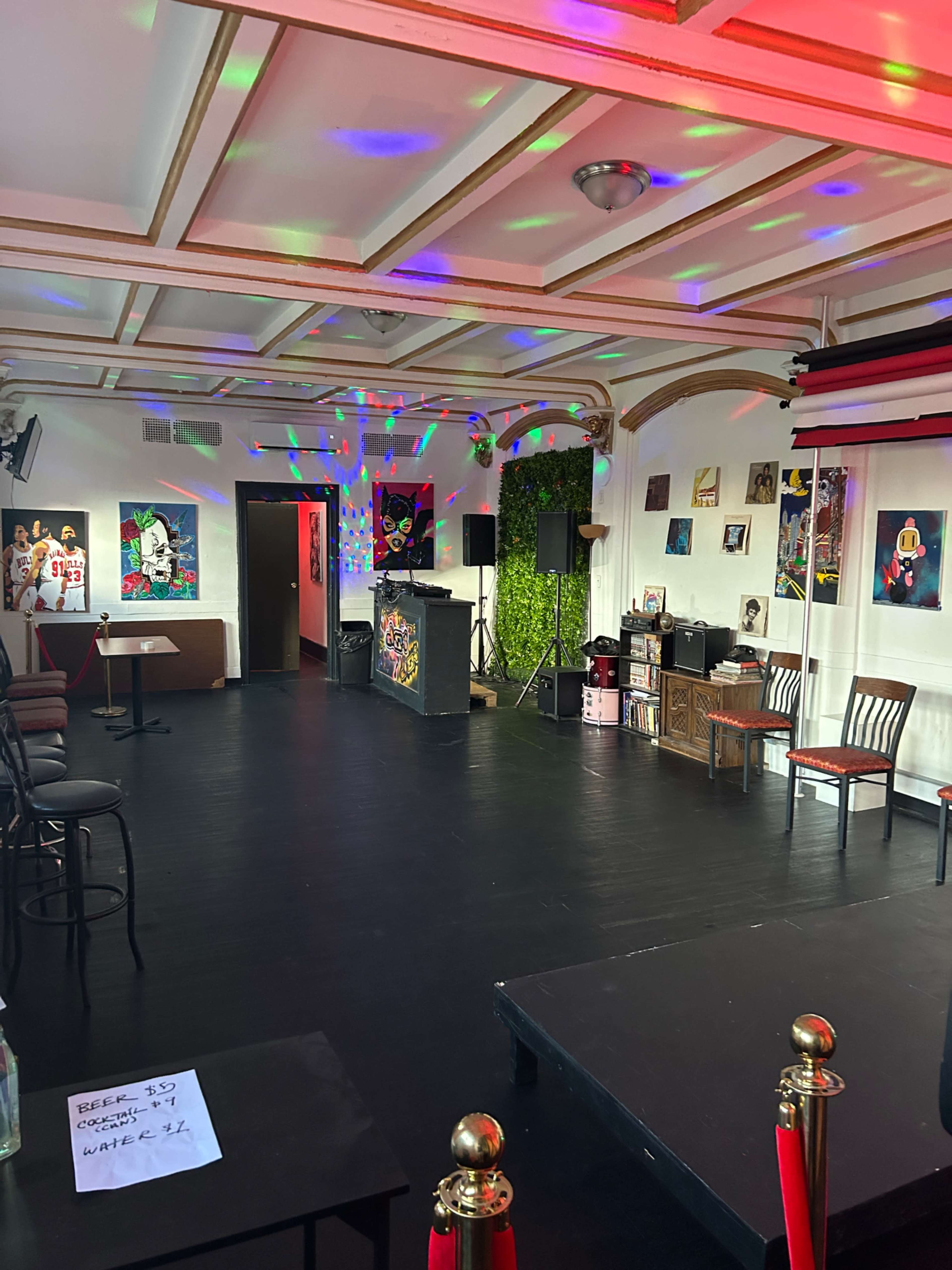 The image shows an empty event space with a DJ booth, colorful lighting, and various artworks on the walls.