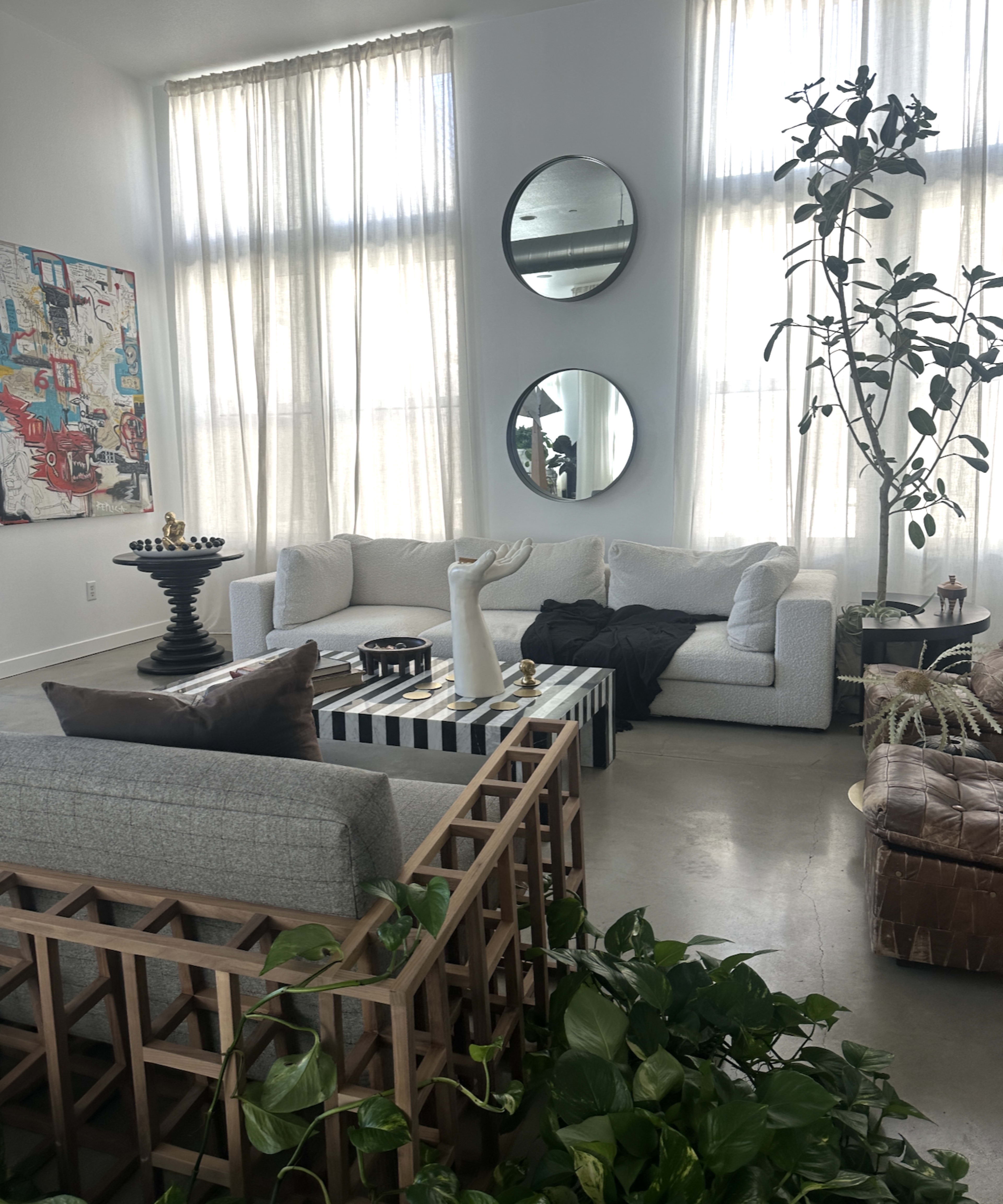 A bright, modern living room features a light-colored sectional sofa, a striped coffee table, and large windows with sheer curtains, alongside decorative plants and wall art.