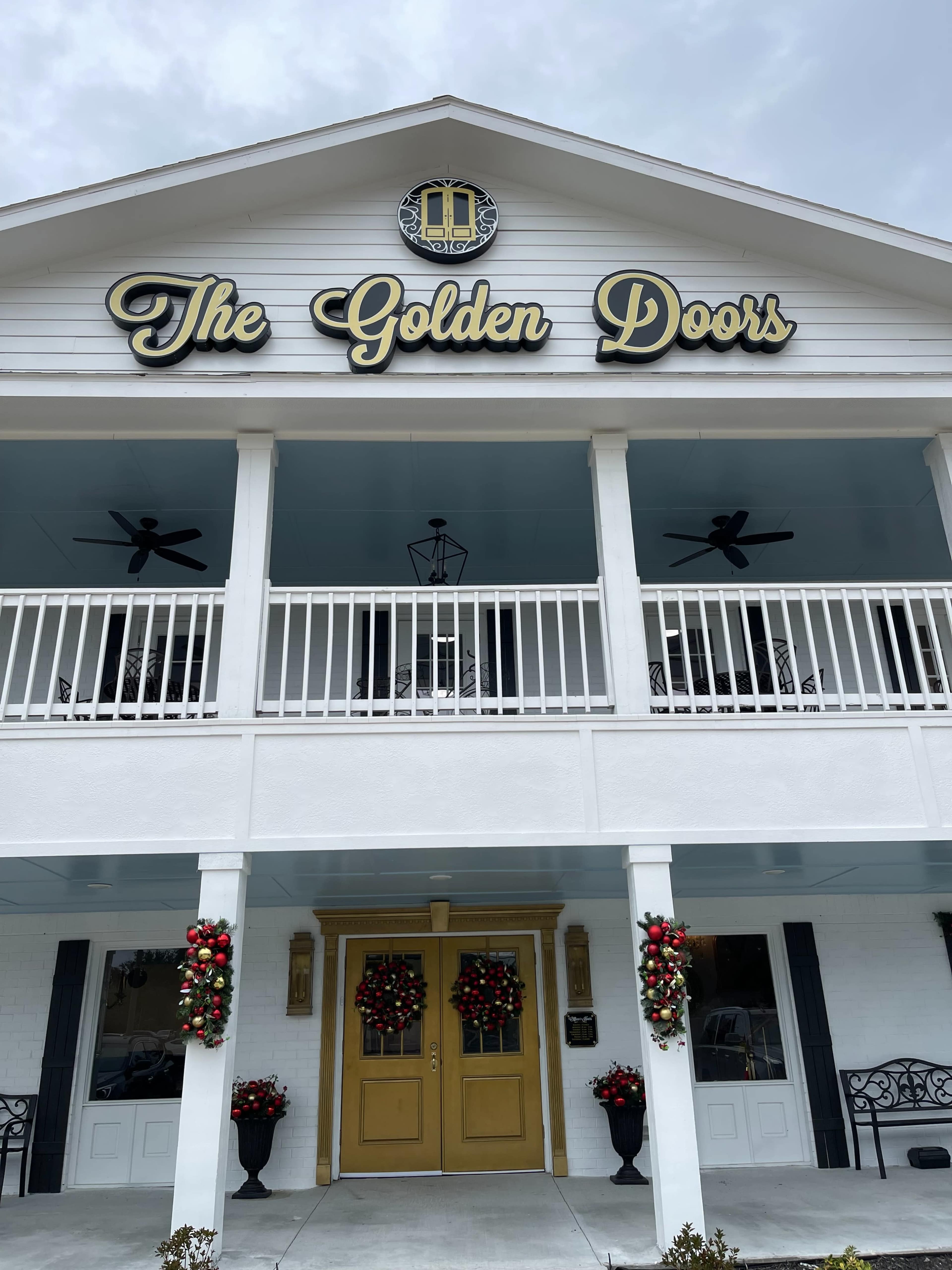 A white building with a porch adorned with fans, featuring the name "The Golden Doors" in gold letters above a welcoming entrance decorated with wreaths.