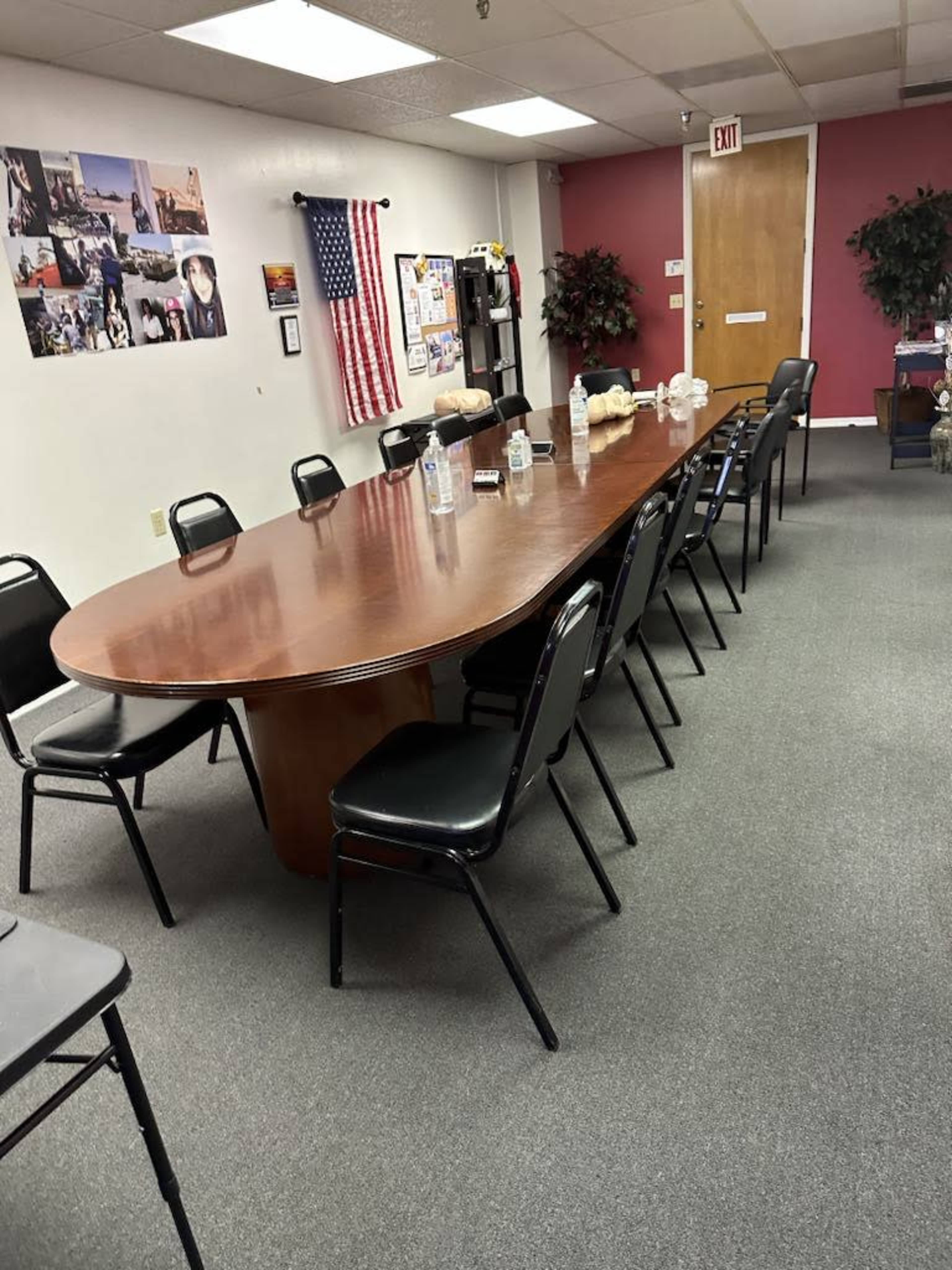 A long, oval wooden conference table is surrounded by black chairs in a room featuring an American flag and various wall decorations.