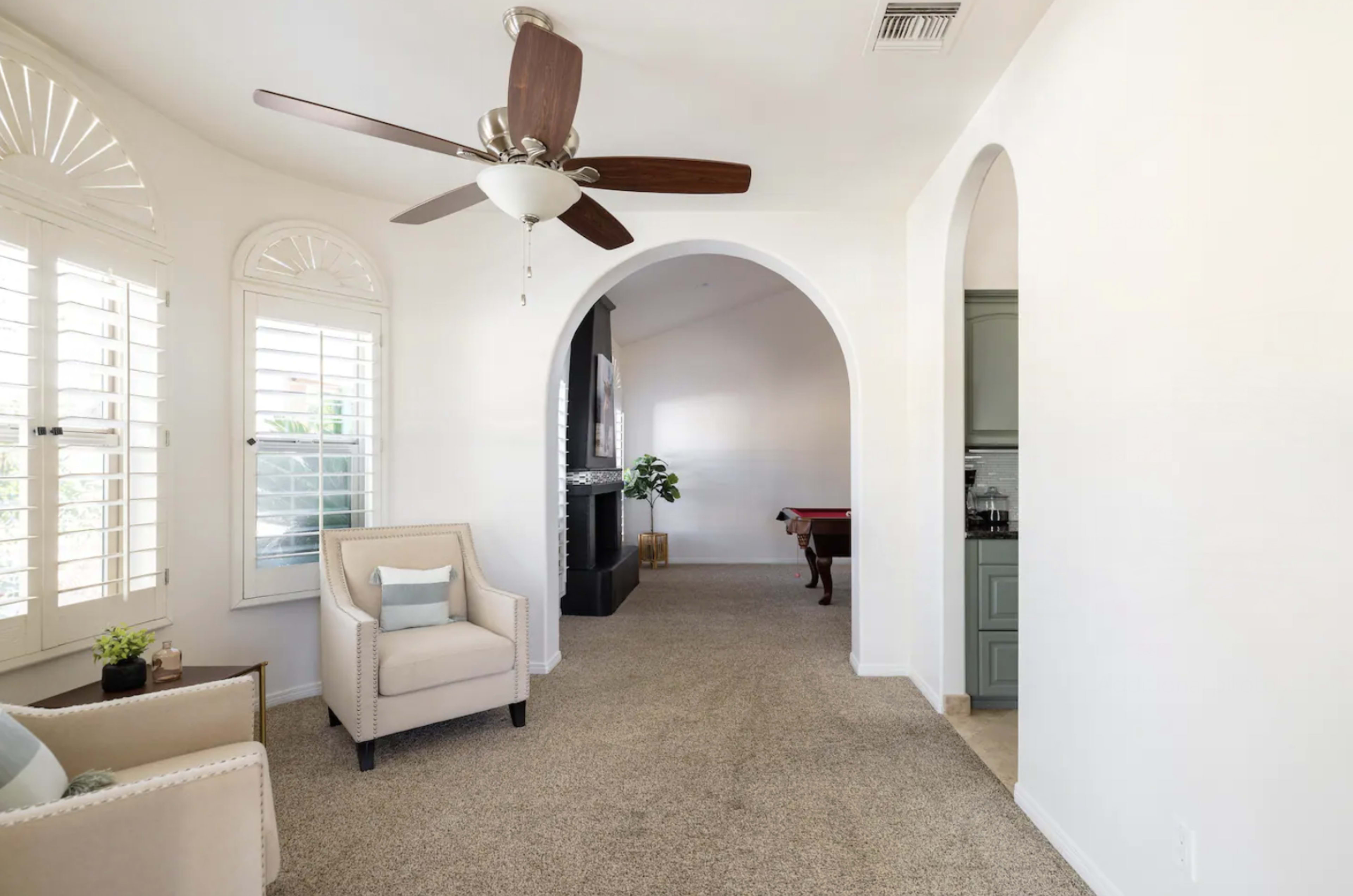 A living area with a ceiling fan, a beige armchair near a window with shutters, and an archway leading to another room.